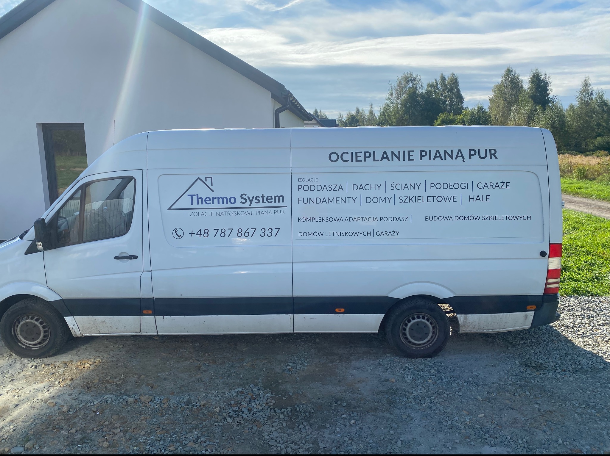 Biały samochód dostawczy z reklamą firmy Thermo System, specjalizującej się w izolacjach natryskowych pianą PUR, zaparkowany na żwirowym podłożu przed budynkiem.