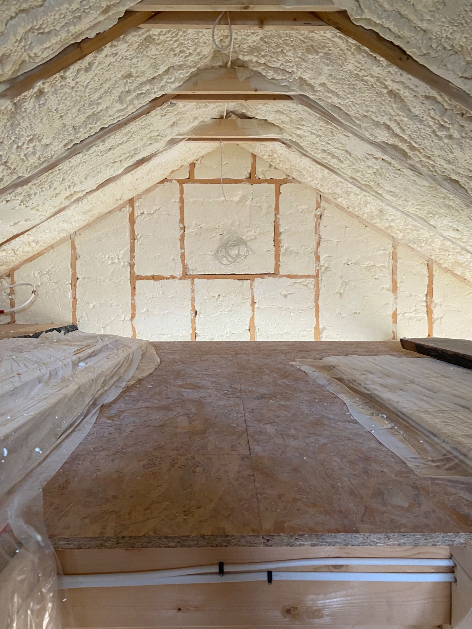 Poddasze z drewnianą konstrukcją pokryte grubą warstwą białej piany izolacyjnej, widoczne fragmenty podłogi zabezpieczone folią.