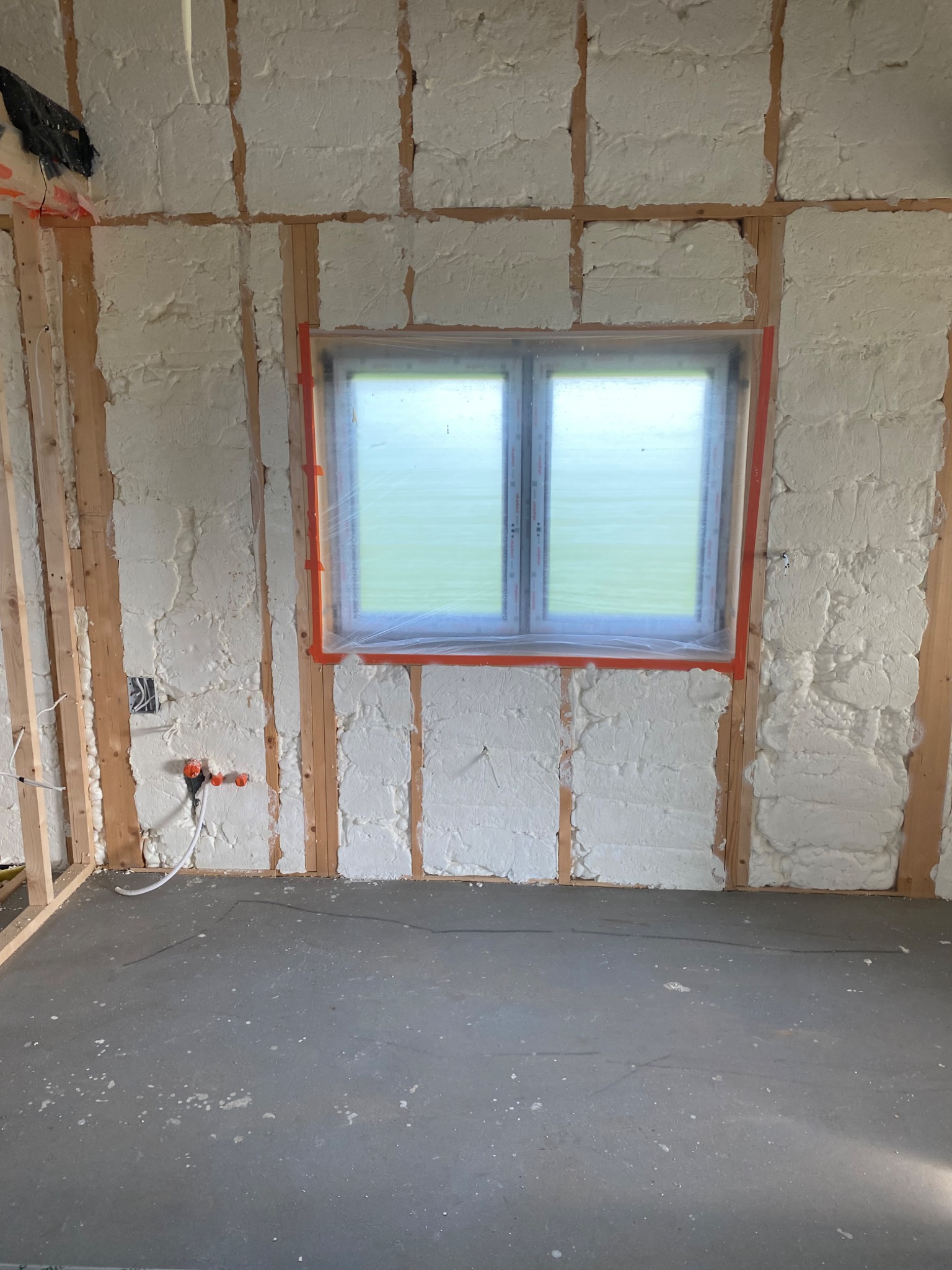 Ściana wewnętrzna w trakcie izolacji, widoczne drewniane belki konstrukcyjne i wypełnienie pianką izolacyjną, okno zabezpieczone folią malarską.