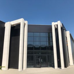 NOWE WNĘTRZE SPÓŁKA Z OGRANICZONĄ ODPOWIEDZIALNOŚCIĄ SPÓŁKA KOMANDYTOWA - Nowoczesny budynek biurowy z czarną elewacją i białymi, wystającymi kolumnami, duże okna z refleksami nieba, otwarte drzwi wejściowe, błękitne niebo w tle.