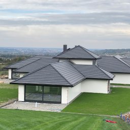 NOWE WNĘTRZE SPÓŁKA Z OGRANICZONĄ ODPOWIEDZIALNOŚCIĄ SPÓŁKA KOMANDYTOWA - Nowoczesny dom jednorodzinny z płaskim dachem pokrytym ciemnoszarą dachówką, białą elewacją i dużymi oknami, widok z góry na tle rozległego krajobrazu miejskiego.