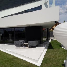 NOWE WNĘTRZE SPÓŁKA Z OGRANICZONĄ ODPOWIEDZIALNOŚCIĄ SPÓŁKA KOMANDYTOWA - Nowoczesny dom z białą elewacją, dużymi oknami i tarasem z meblami ogrodowymi przykrytymi pokrowcami, widoczna antena satelitarna na balkonie.
