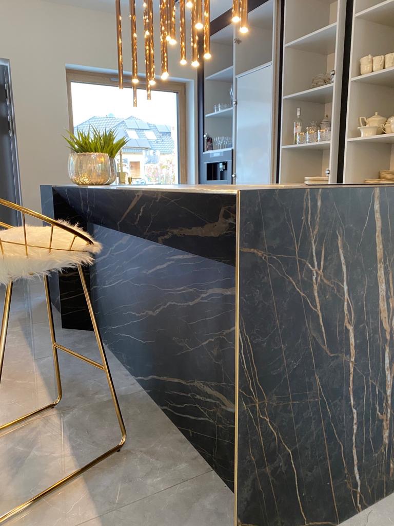 Nowoczesna wyspa kuchenna z czarnym blatem imitującym marmur ze złotymi żyłami, złoty hoker z białym futrzanym siedziskiem, szara podłoga, oświetlenie wiszące w kolorze złota i białe szafki w tle.