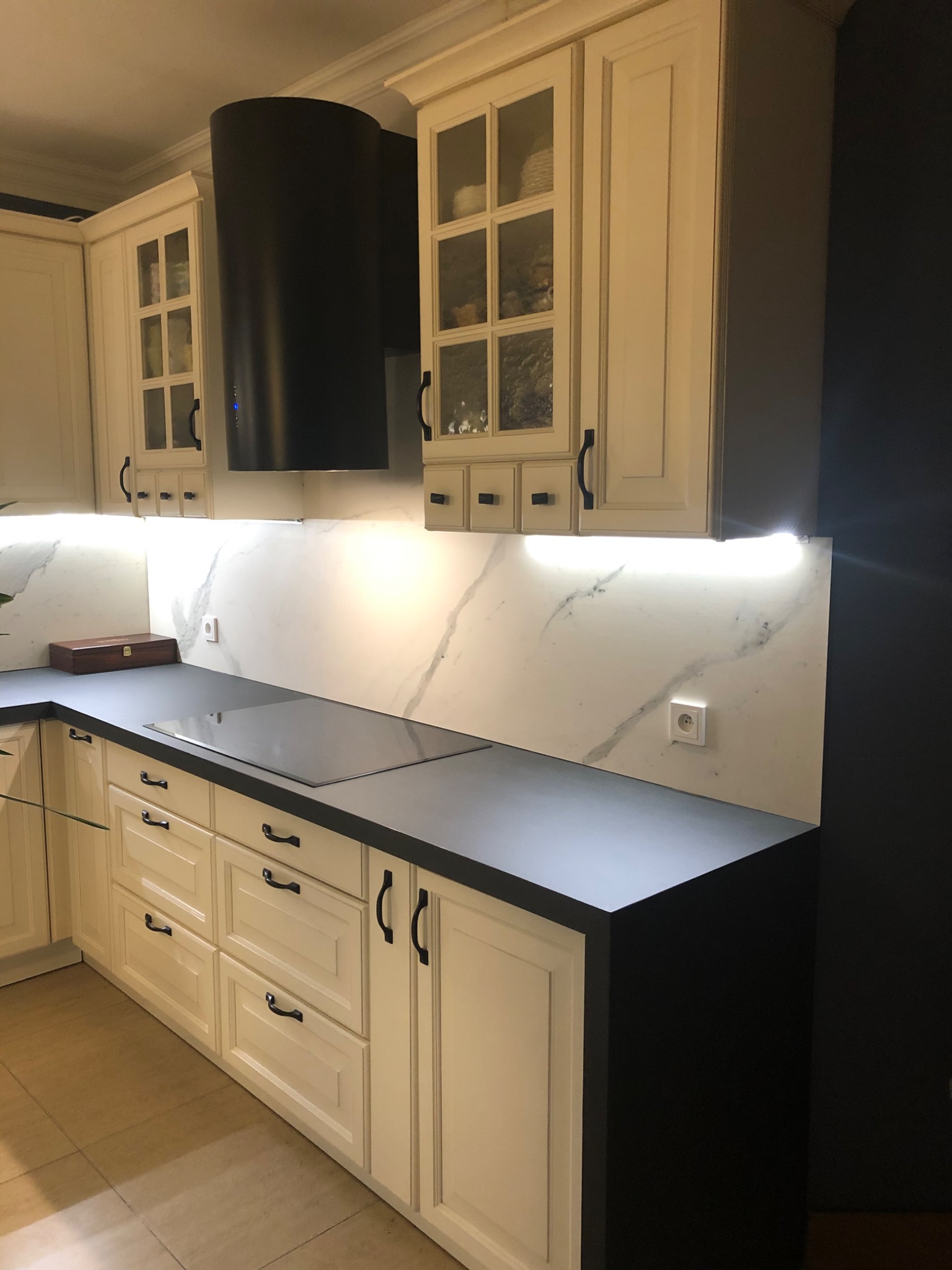 Kremowa zabudowa kuchenna z czarnym blatem i okapem, marmurowy panel nad blatem z oświetleniem LED, czarne uchwyty szafek, widoczna płyta indukcyjna i drewniana szkatułka.