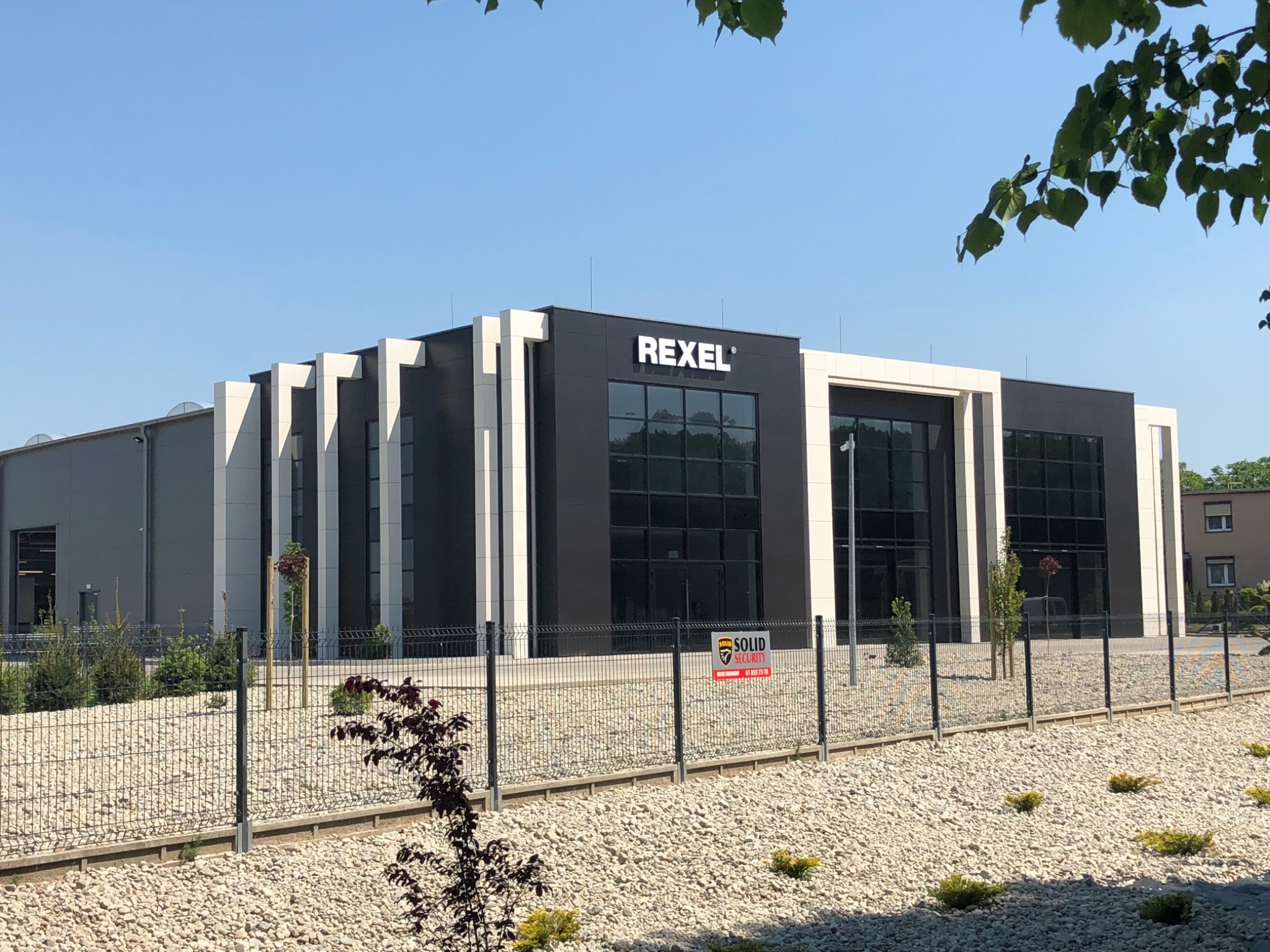 Nowoczesny budynek biurowy Rexel z czarno-białą elewacją, dużymi oknami i minimalistycznym designem, otoczony żwirowym terenem i ogrodzeniem z siatki.
