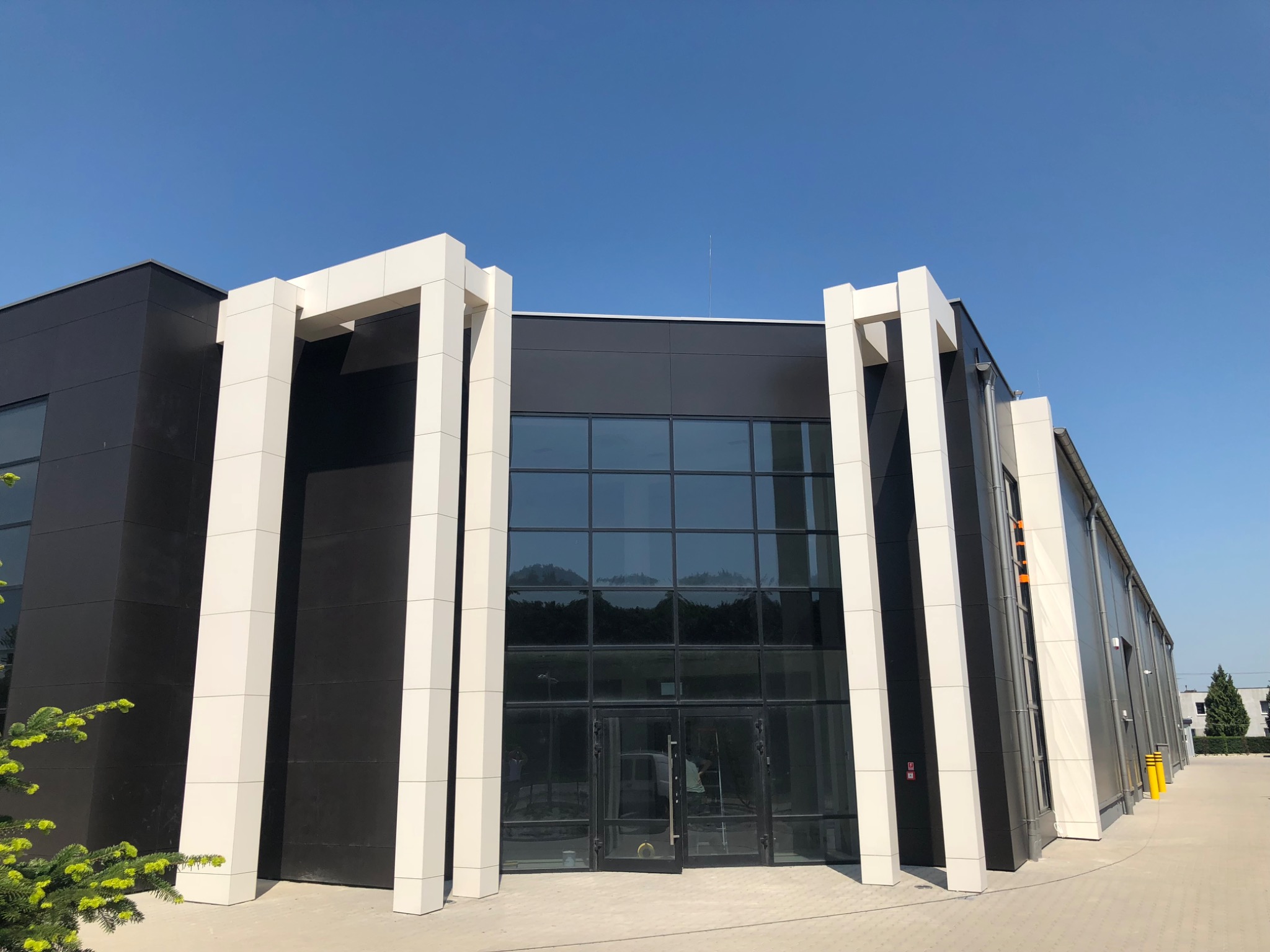 Nowoczesny budynek biurowy z czarną elewacją i białymi, wystającymi kolumnami, duże okna z refleksami nieba, otwarte drzwi wejściowe, błękitne niebo w tle.