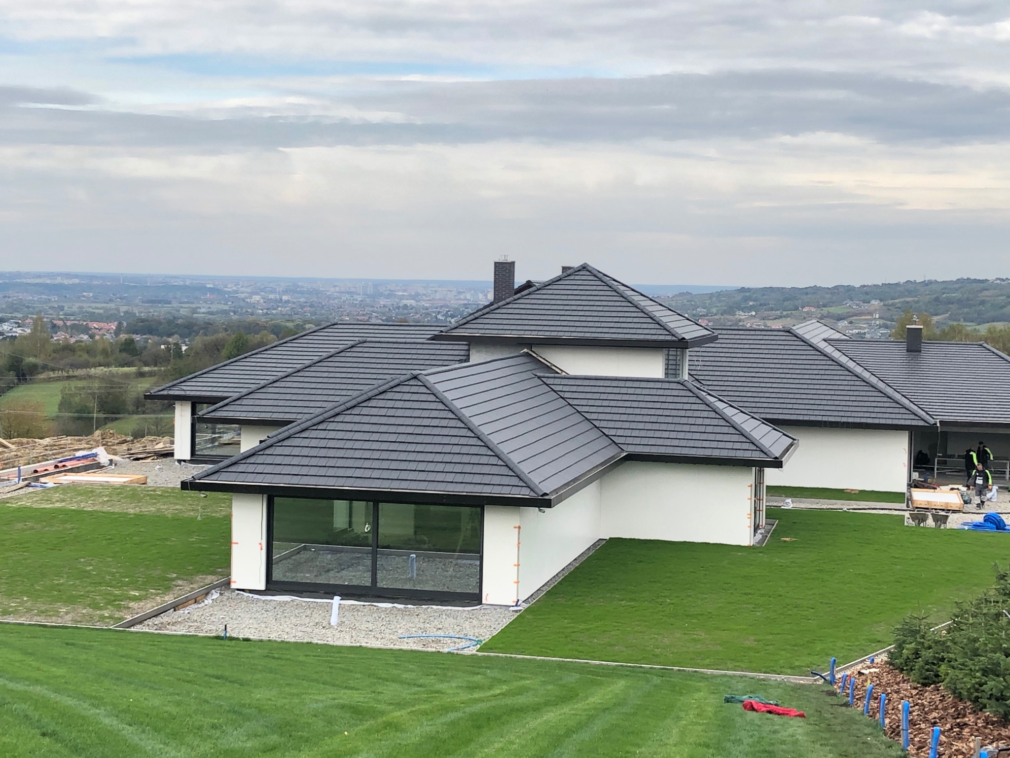 Nowoczesny dom jednorodzinny z płaskim dachem pokrytym ciemnoszarą dachówką, białą elewacją i dużymi oknami, widok z góry na tle rozległego krajobrazu miejskiego.