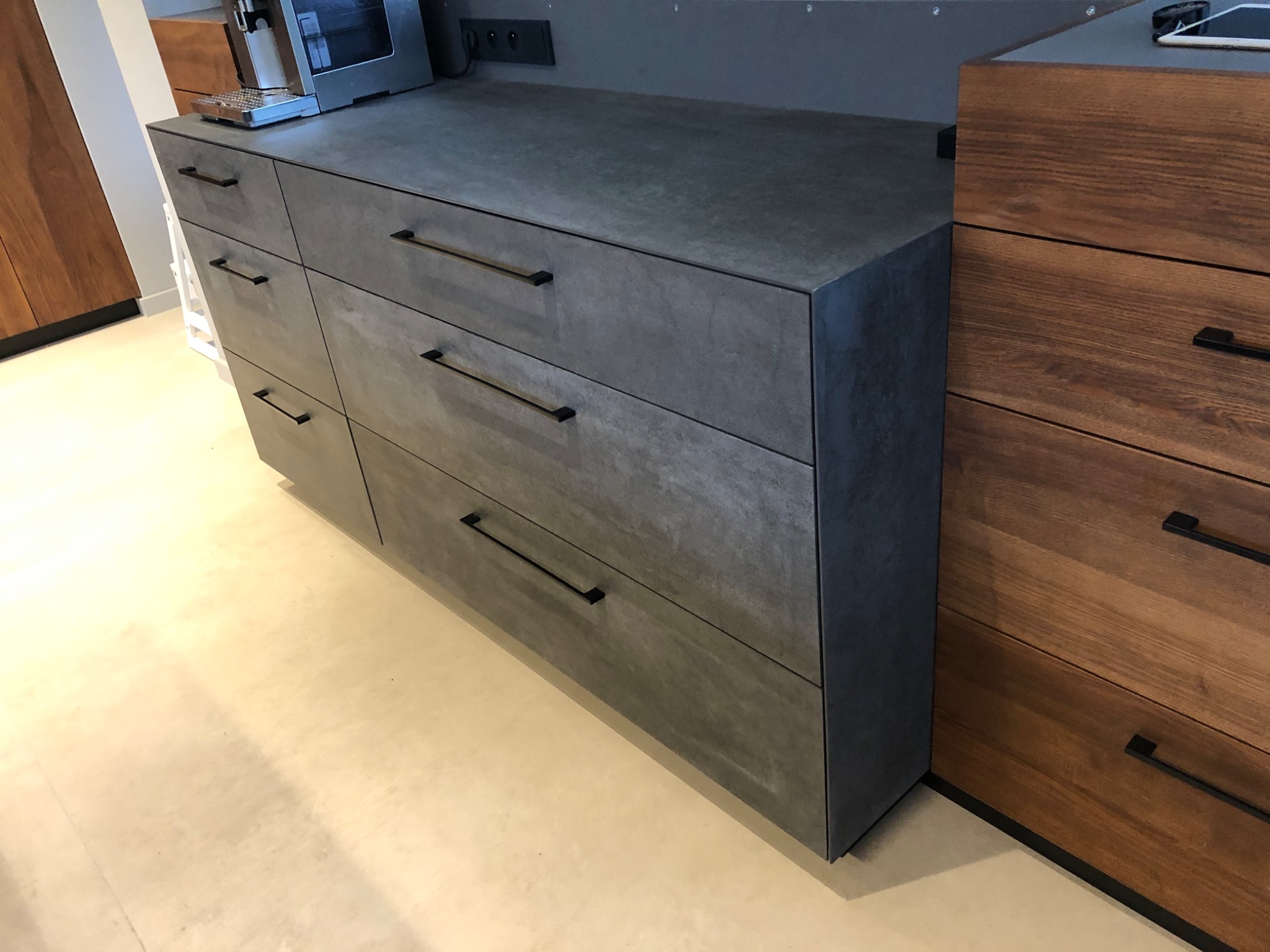 Szare, betonowe szafki z czarnymi, prostokątnymi uchwytami, sąsiadujące z drewnianymi szafkami z widocznym usłojeniem. Na blacie szafek stoi ekspres do kawy i ładowarka. Podłoga w jasnym odcieniu...