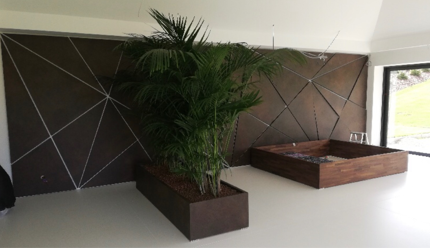 Nowoczesne wnętrze z ciemną ścianą ozdobioną geometrycznym wzorem z jasnych linii, donicą z palmą areka i drewnianą konstrukcją przypominającą podest lub siedzisko.