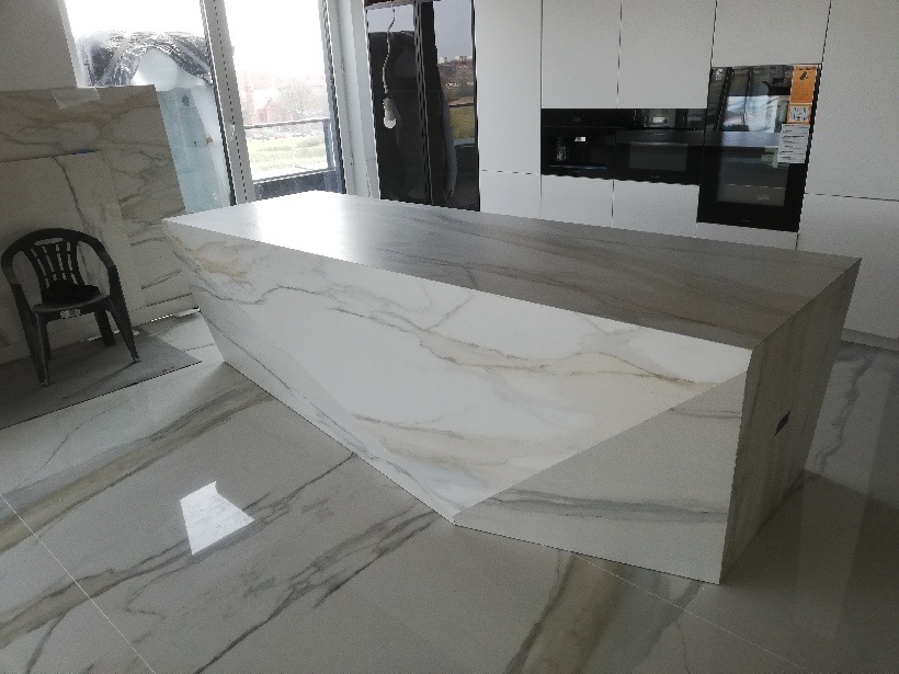 Wykończona wyspa kuchenna z białego marmuru z szarymi żyłami, dopasowana do marmurowej podłogi, na tle białych szafek kuchennych z wbudowanym piekarnikiem.