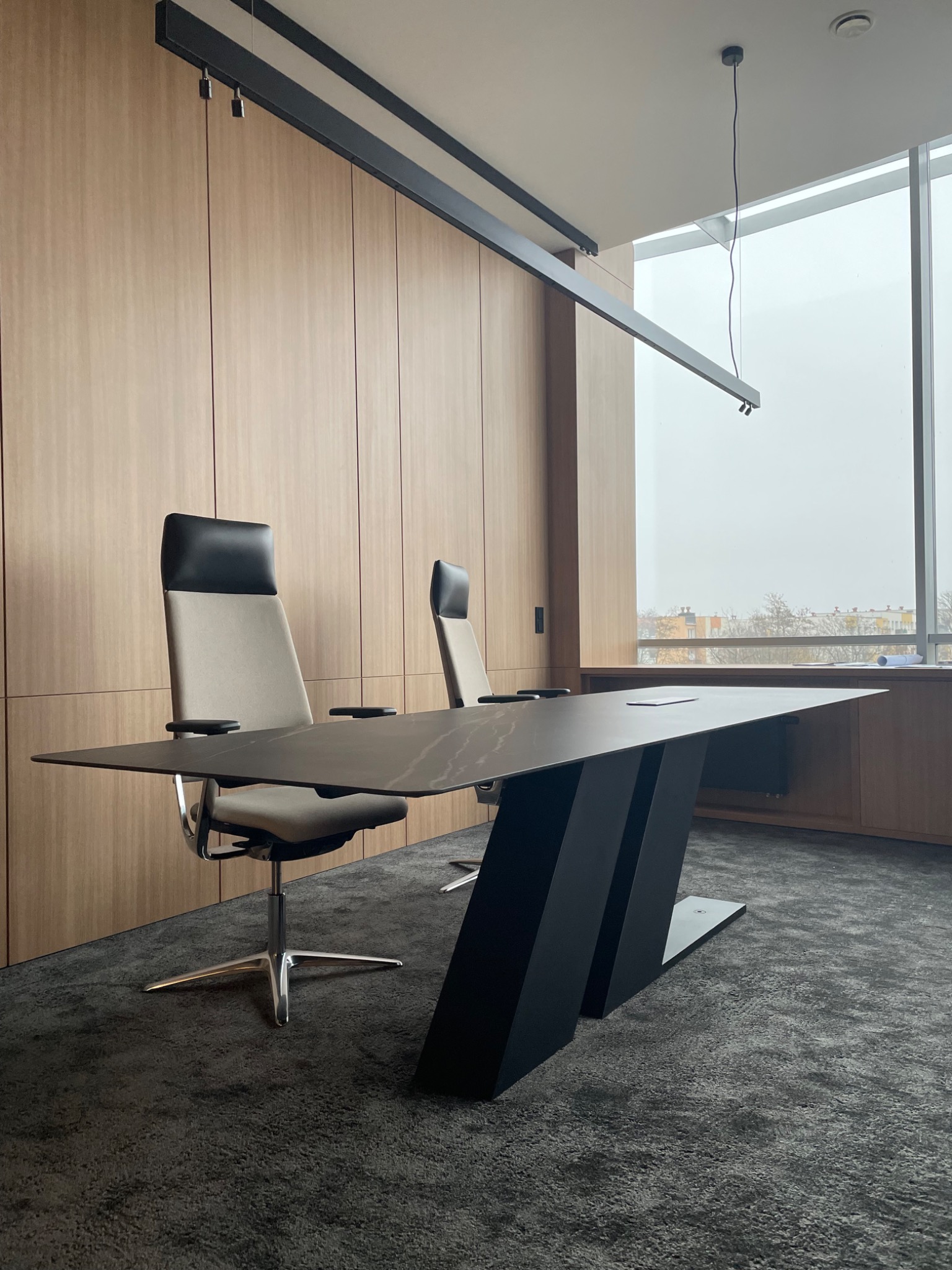 Nowoczesne biuro: Duży stół konferencyjny z czarnym blatem i geometryczną podstawą, dwie ergonomiczne krzesła z szarym siedziskiem i czarnym zagłówkiem, drewniana okładzina ścienna oraz duże okno...
