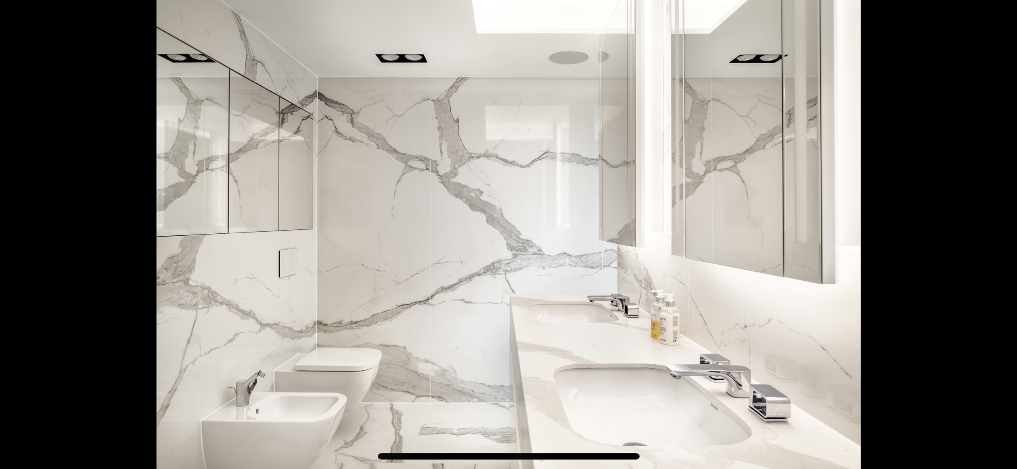 Nowoczesna łazienka wykończona białym marmurem z szarymi żyłami, wyposażona w dwie umywalki nablatowe z chromowanymi bateriami, bidet i toaletę w minimalistycznym stylu, oświetlona punktowymi...