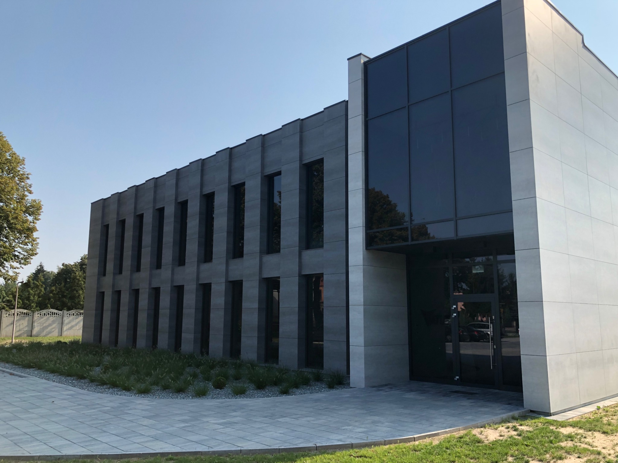 Nowoczesny budynek biurowy z elewacją z szarej płyty, duże okna i przeszklone wejście, otoczony zielenią i chodnikiem z kostki brukowej.