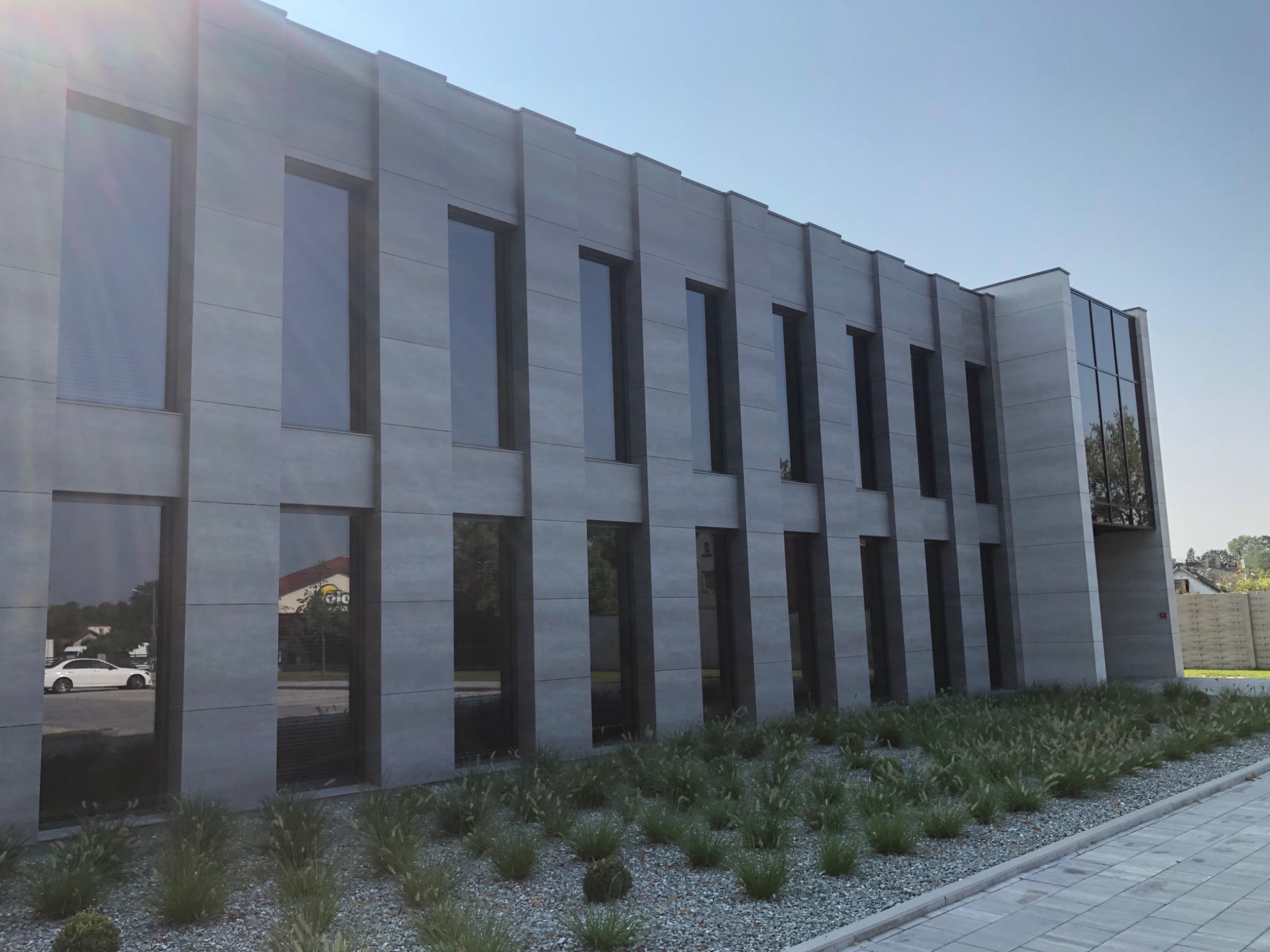 Nowoczesny budynek biurowy z szarą elewacją panelową i dużymi oknami, w których odbija się otoczenie. Na pierwszym planie dekoracyjny żwirowy ogródek z trawami ozdobnymi.