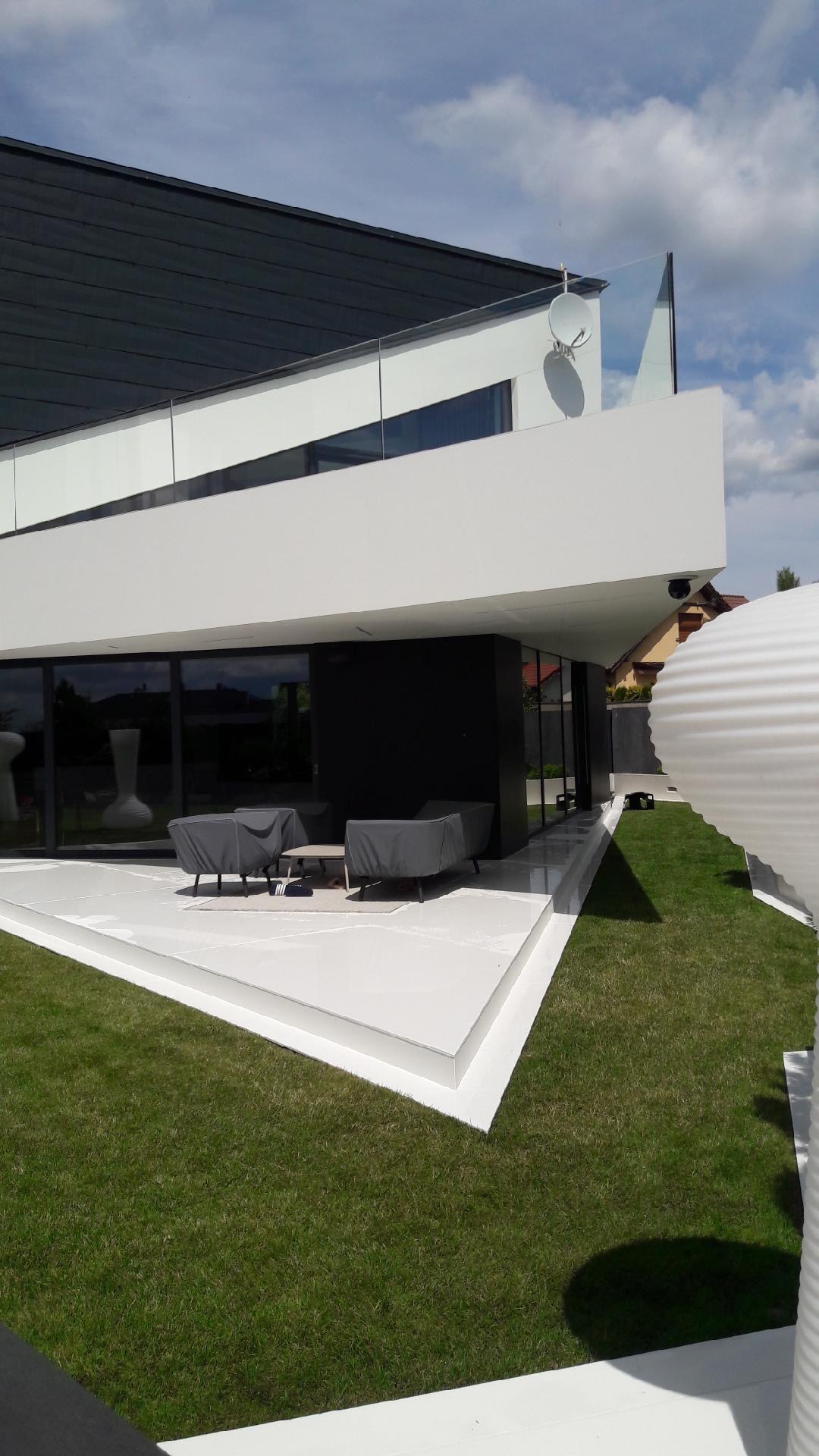 Nowoczesny dom z białą elewacją, dużymi oknami i tarasem z meblami ogrodowymi przykrytymi pokrowcami, widoczna antena satelitarna na balkonie.