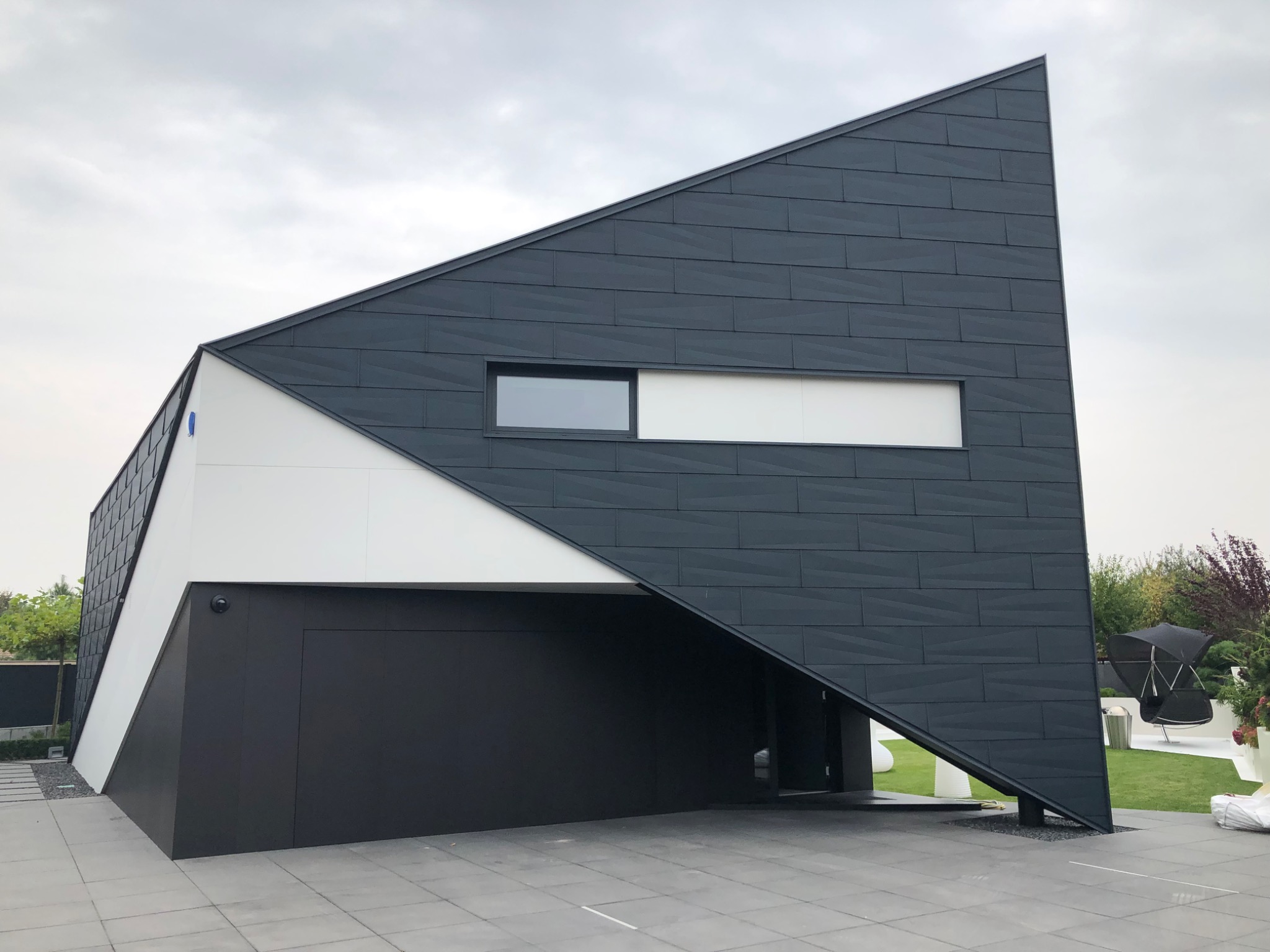 Nowoczesny dom jednorodzinny z elewacją w kolorze antracytu, o nietypowej, trójkątnej bryle, z białymi akcentami i dużymi oknami, otoczony zadbanym ogrodem.