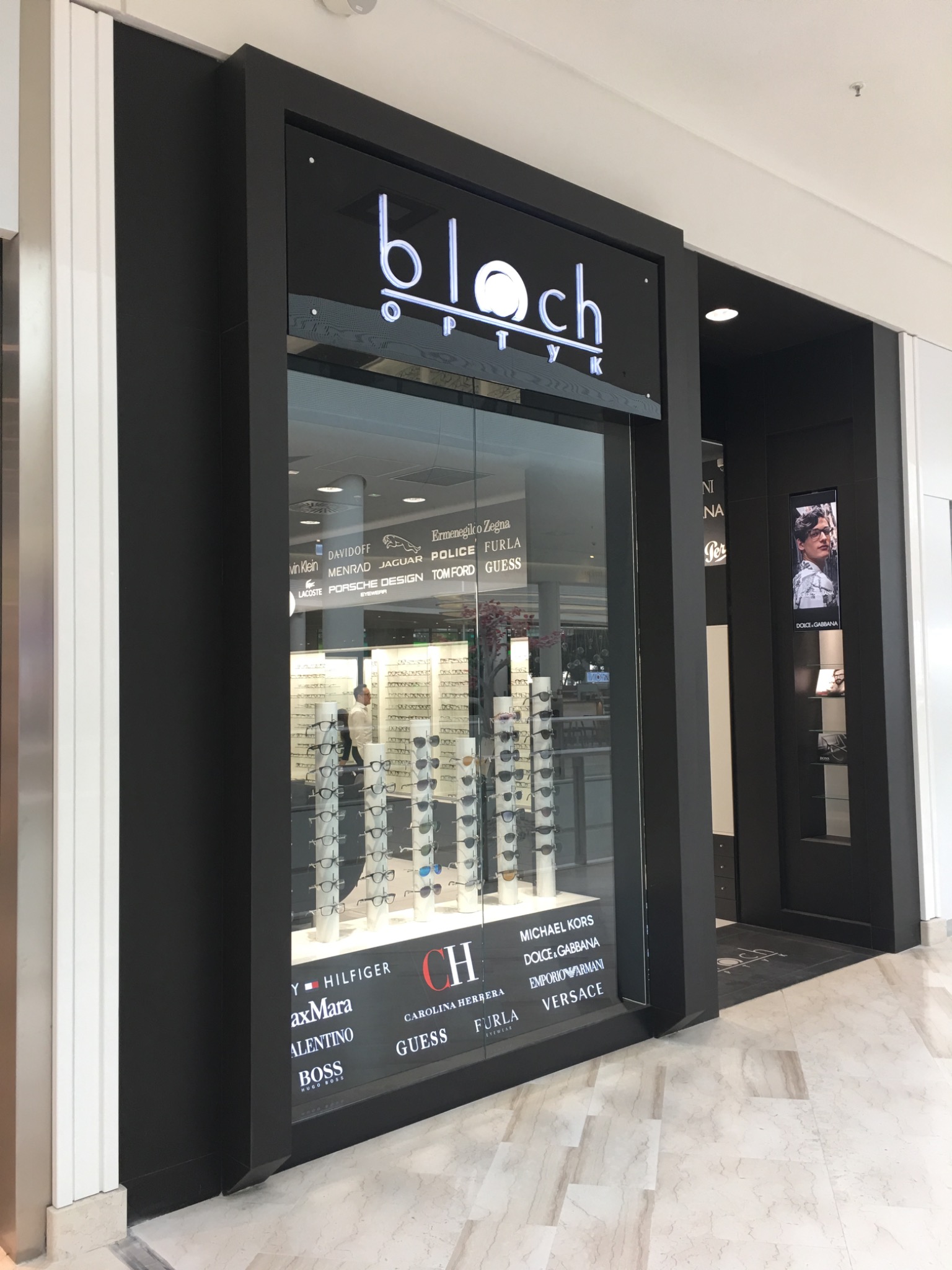 Elegancka witryna salonu optycznego 'Bloch Optyk' z ekspozycją okularów różnych marek, takich jak Dolce & Gabbana, Versace, Furla, umieszczona w nowoczesnym centrum handlowym z marmurową podłogą.