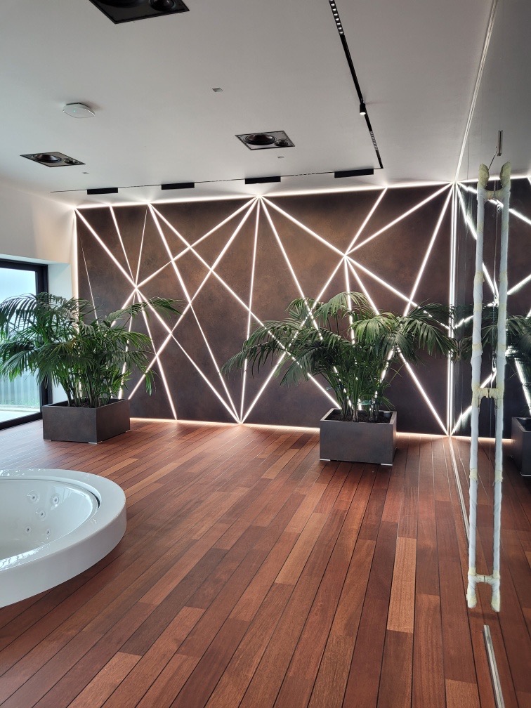 Nowoczesna łazienka z drewnianą podłogą, okrągłą wanną z hydromasażem i ścianą dekoracyjną z podświetlanymi geometrycznymi wzorami oraz roślinami w donicach.
