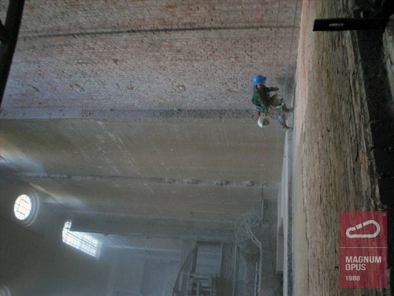 Pracownik w kasku i uprzęży, zawieszony na linie, czyści ceglaną ścianę budynku od góry, widok z dołu na wnętrze z oknami.