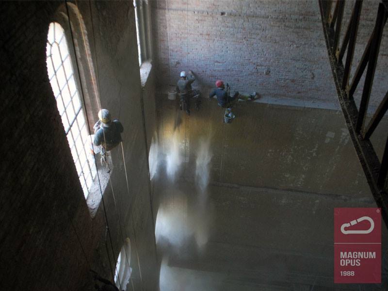 Trzech alpinistów podczas prac wysokościowych przy renowacji ceglanej ściany budynku z dużymi oknami łukowymi, widoczne liny, uprzęże i wiaderka z zaprawą.