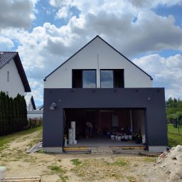 SIDO- USŁUGI OGÓLNOBUDOWLANE SYLWESTER GAWŁOWSKI - Dwukondygnacyjny dom z szarą elewacją w trakcie malowania. Widoczne puste okna na górze i otwarty garaż z farbami na dole. Prace w toku.