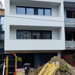 SIDO- USŁUGI OGÓLNOBUDOWLANE SYLWESTER GAWŁOWSKI - Nowoczesny, dwupiętrowy budynek z białą elewacją i czarnymi oknami, w trakcie prac wykończeniowych. Widoczne materiały budowlane i pracownik w odblaskowej kamizelce.