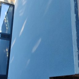 SIDO- USŁUGI OGÓLNOBUDOWLANE SYLWESTER GAWŁOWSKI - Ściana budynku po tynkowaniu, pomalowana na niebiesko, z widocznymi refleksami światła. W tle okno i fragment niezakończonej elewacji z tablicą informacyjną budowy.