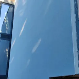 Ściana budynku po tynkowaniu, pomalowana na niebiesko, z widocznymi refleksami światła. W tle okno i fragment niezakończonej elewacji z tablicą informacyjną budowy.