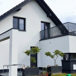 SIDO- USŁUGI OGÓLNOBUDOWLANE SYLWESTER GAWŁOWSKI - Nowoczesny dom z białą elewacją i czarnym dachem, balkon z metalową balustradą, widok z boku, zieleń w tle, pracownik w tle po lewej.