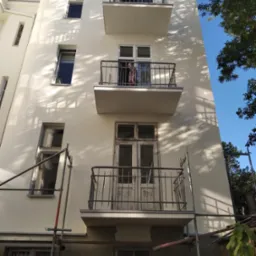 Budynek wielorodzinny z białymi elewacjami i balkonami, widoczne rusztowania i ślady prac wykończeniowych na elewacji.