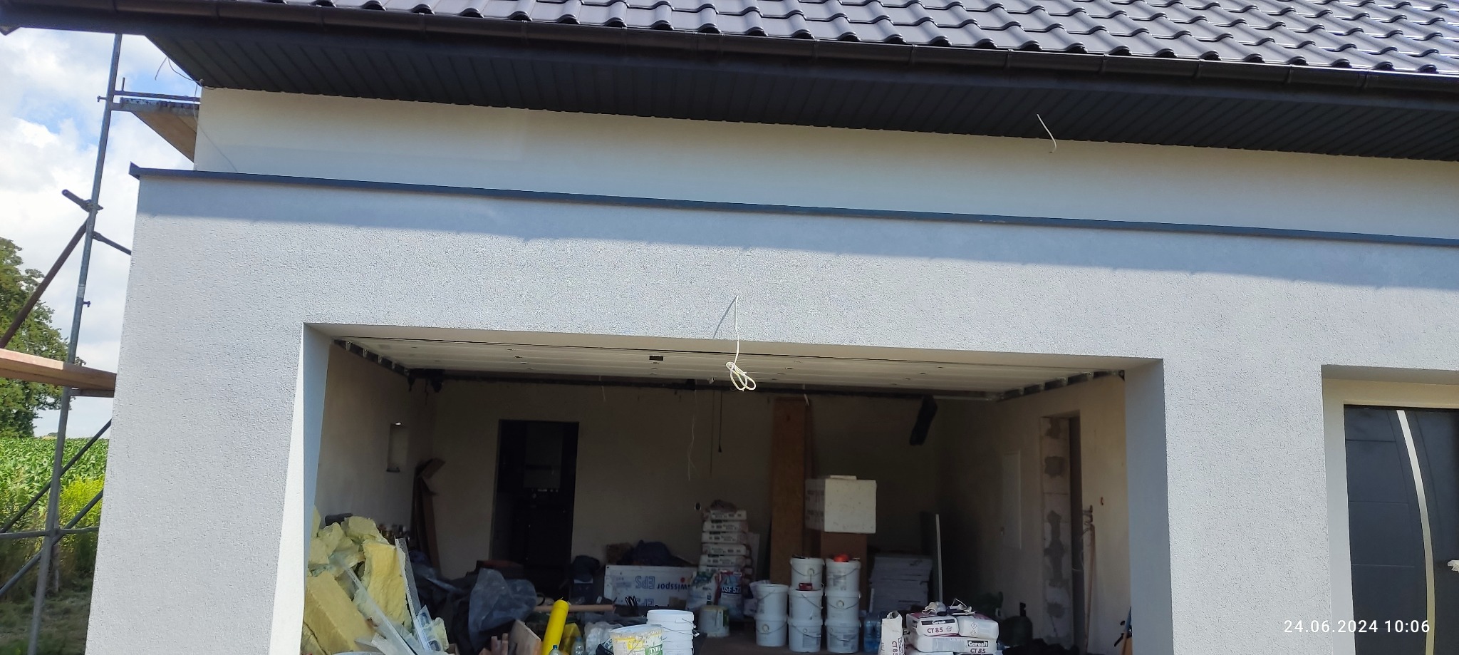 Dom w trakcie prac wykończeniowych elewacji, widoczne rusztowanie, ocieplenie i materiały budowlane w otwartym garażu, zarysowana faktura tynku zewnętrznego.