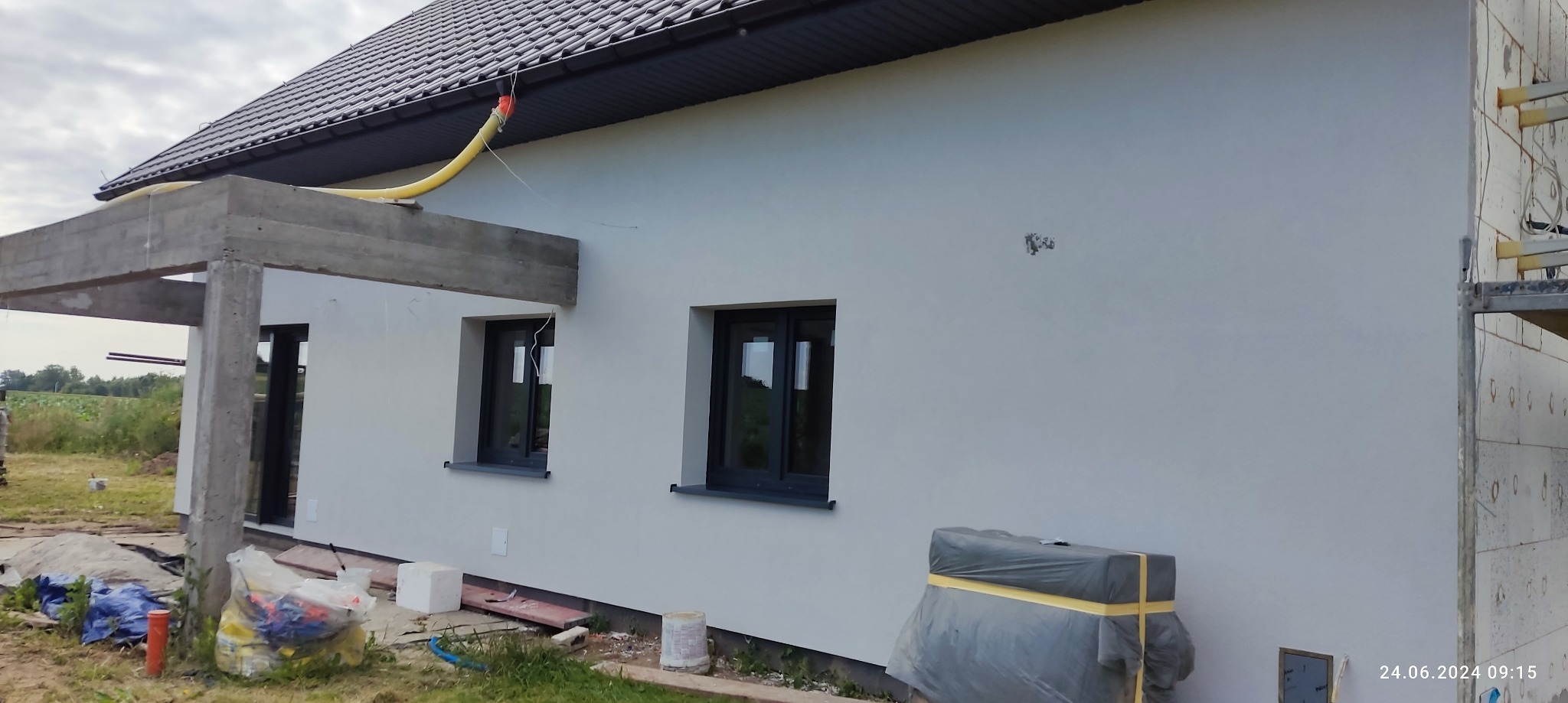 Świeżo otynkowana elewacja domu jednorodzinnego z czarnymi oknami, widoczne ślady po mocowaniu rusztowania oraz żółty wąż wentylacyjny pod dachem.