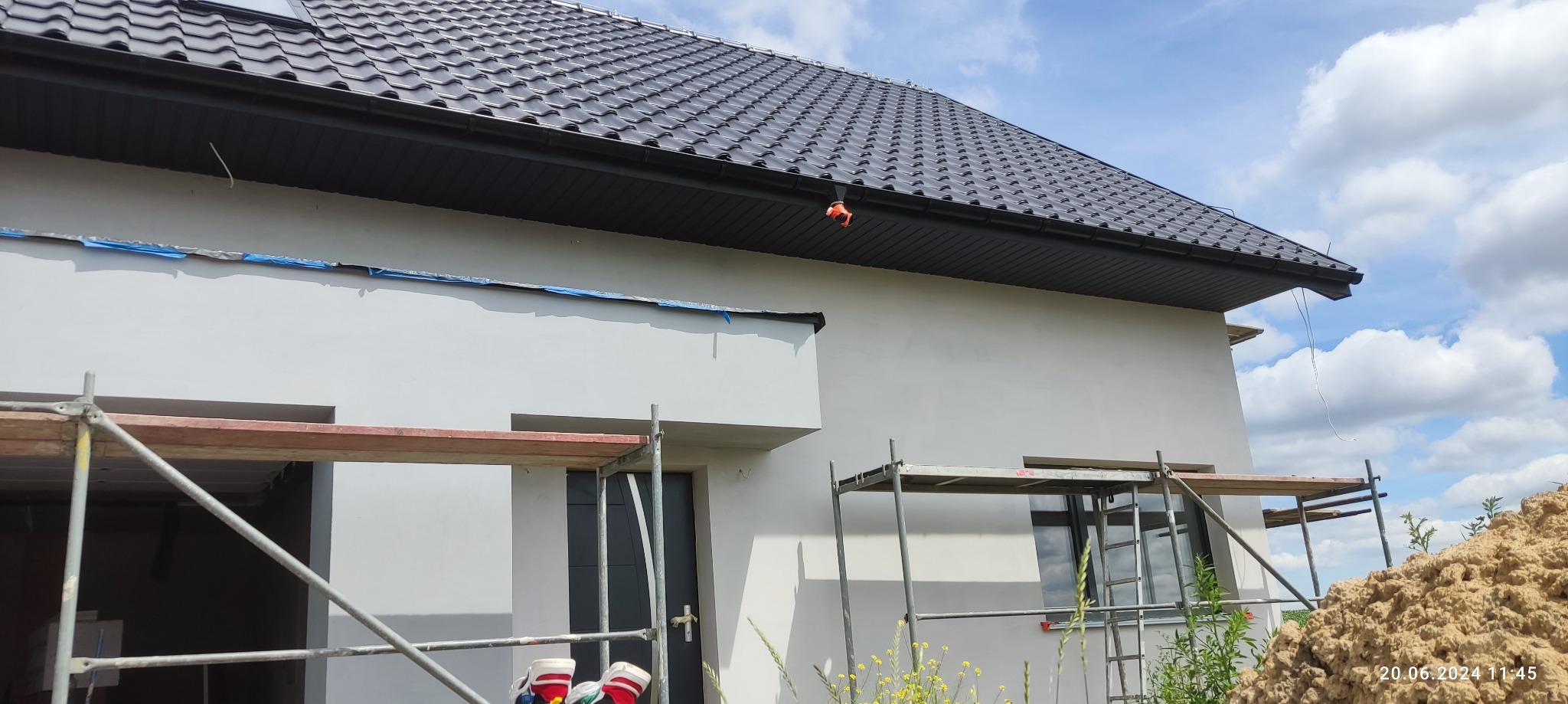 Świeżo otynkowana elewacja domu jednorodzinnego w trakcie prac wykończeniowych, widoczne rusztowania i zabezpieczenia krawędzi taśmą malarską.