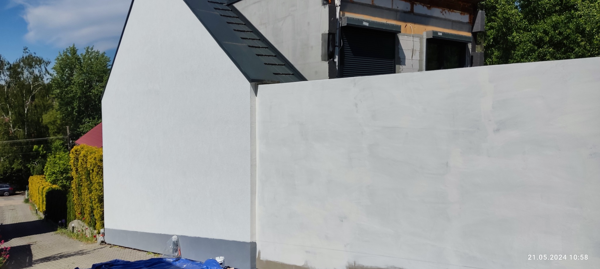 Świeżo pomalowana, biała elewacja domu jednorodzinnego w trakcie prac wykończeniowych, widoczne ślady po wałku i niedomalowane fragmenty, z boku fragment chodnika i zieleni.
