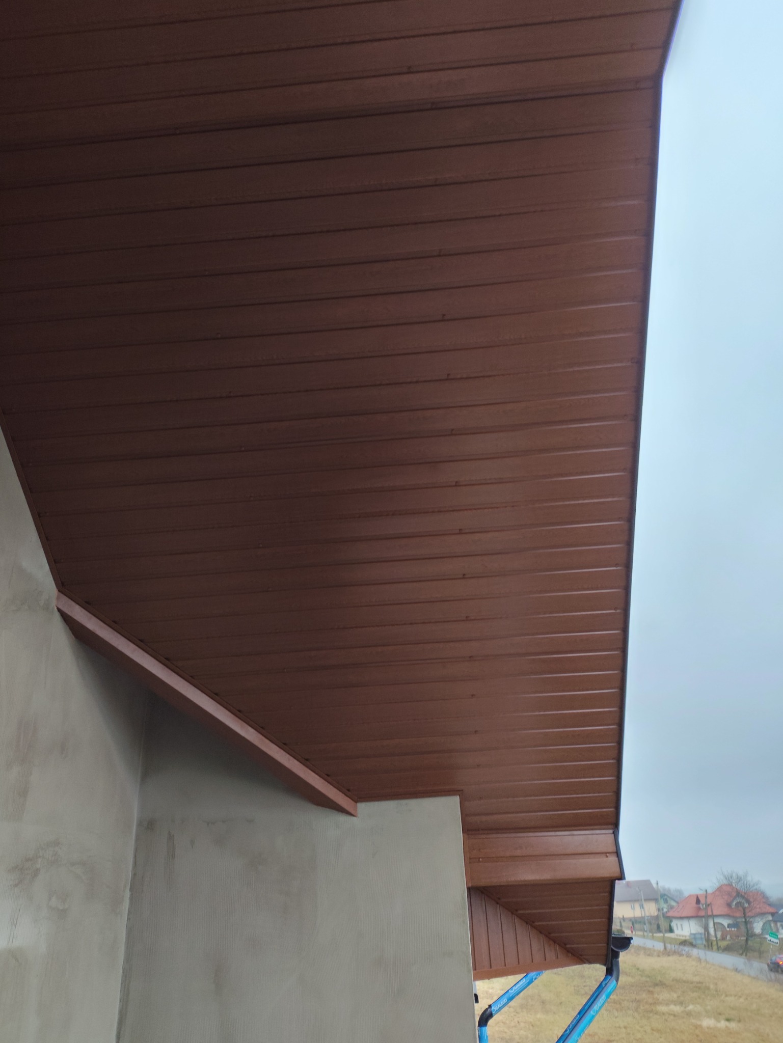 Widok z dołu na brązową podbitkę dachową z PCV, zamontowaną na budynku ze świeżo otynkowaną ścianą zewnętrzną, w tle pochmurne niebo i zabudowania.