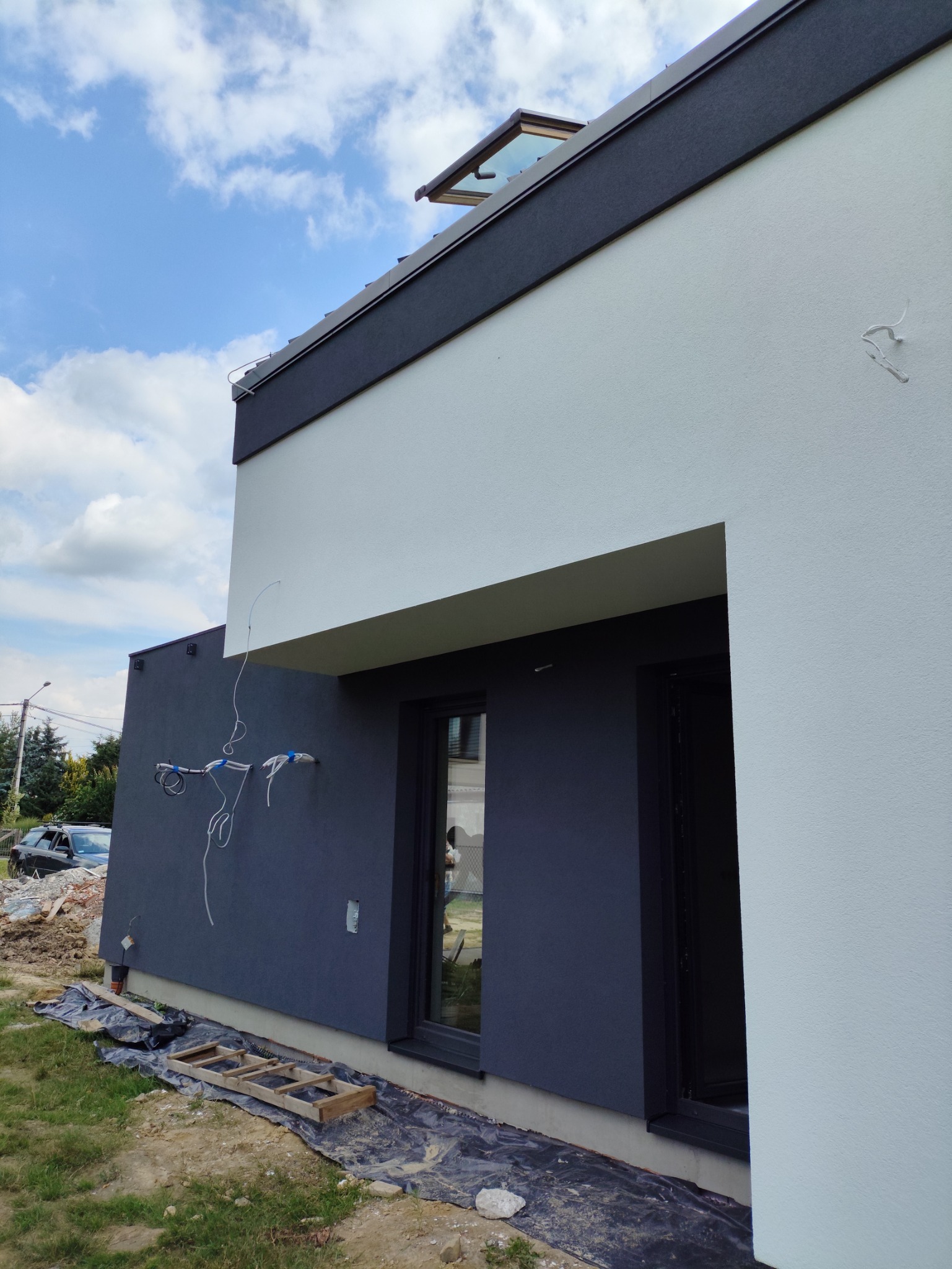 Dwukolorowa elewacja domu z widocznymi przewodami na ścianie i otwartym oknem dachowym. Częściowo ukończona praca, widoczne zabezpieczenia.