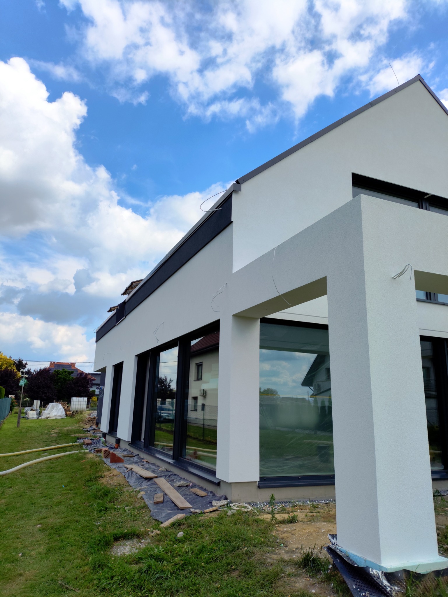 Nowoczesny dom z białą elewacją i dużymi oknami, widok z zewnątrz, na trawniku widoczne materiały budowlane. Dom w stylu minimalistycznym, błękitne niebo z chmurami w tle.