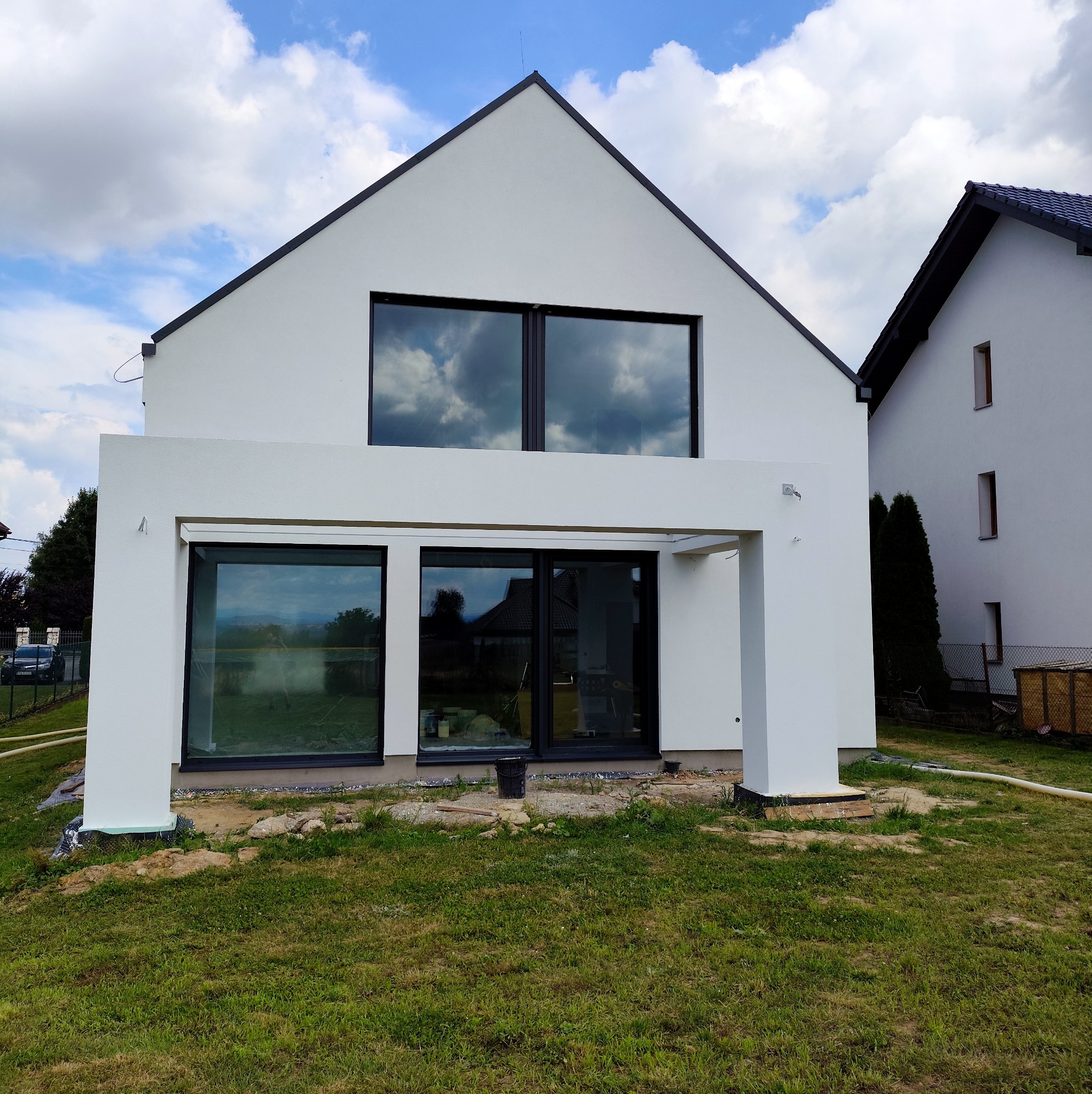 Nowoczesny dom z białą elewacją i dużymi oknami, w których odbijają się chmury. Widoczne ślady prac wykończeniowych na trawniku. Minimalistyczny design.