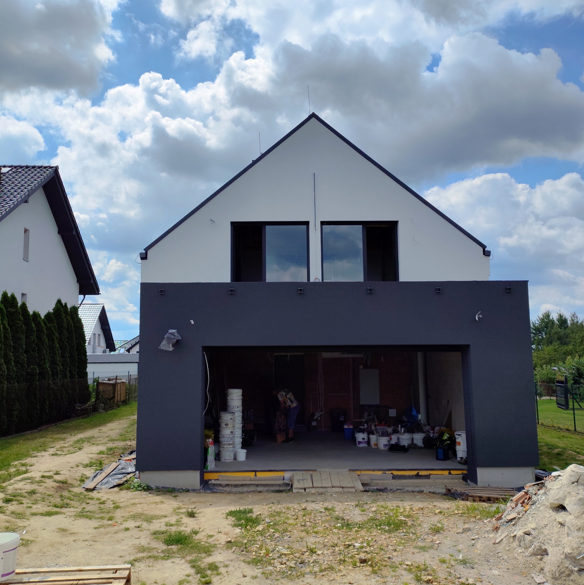 Dwukondygnacyjny dom z szarą elewacją w trakcie malowania. Widoczne puste okna na górze i otwarty garaż z farbami na dole. Prace w toku.