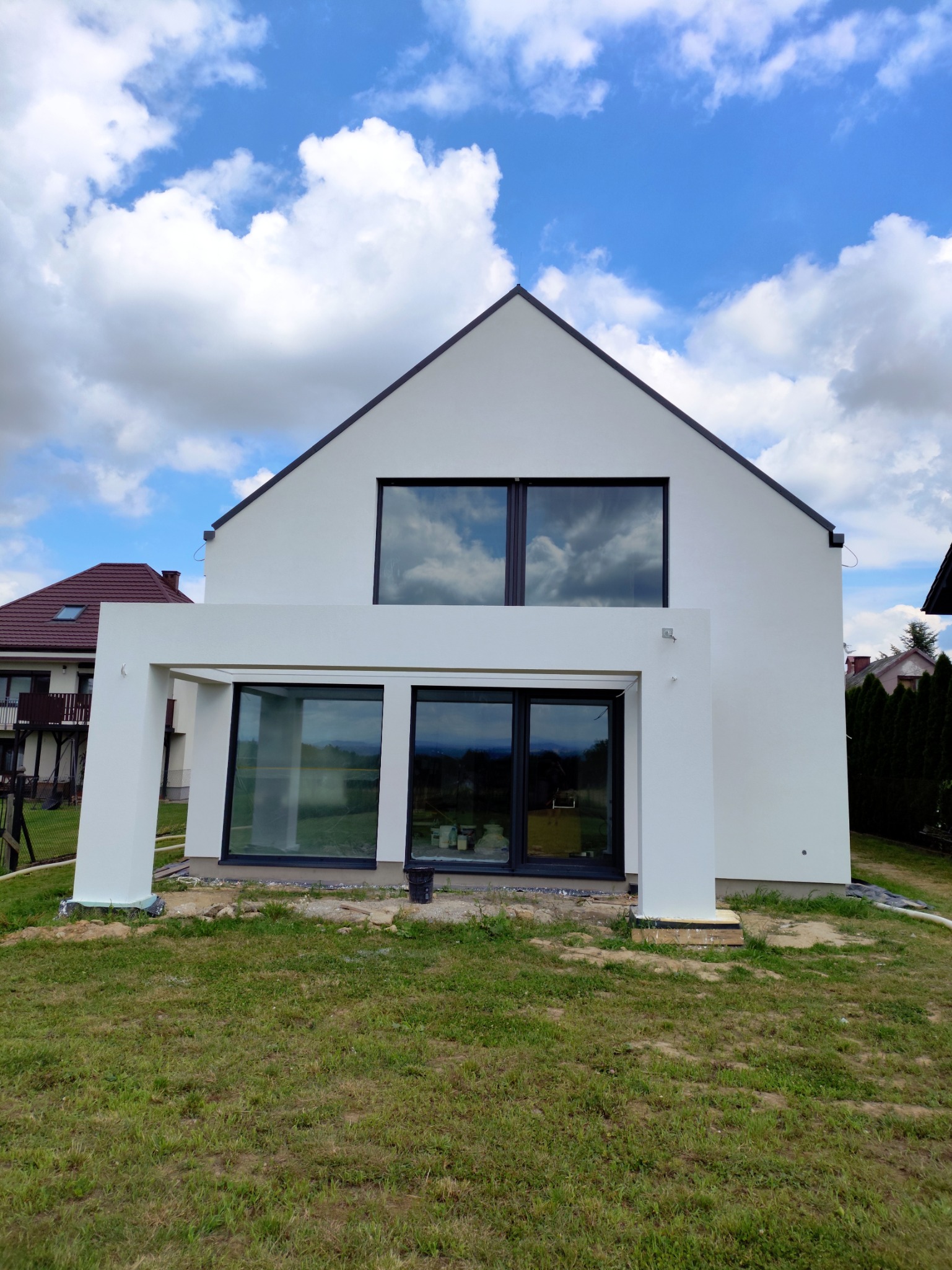 Biały, nowoczesny dom jednorodzinny z dużymi oknami, na tle błękitnego nieba z chmurami. Widoczne elementy elewacji i fragment ogrodu.