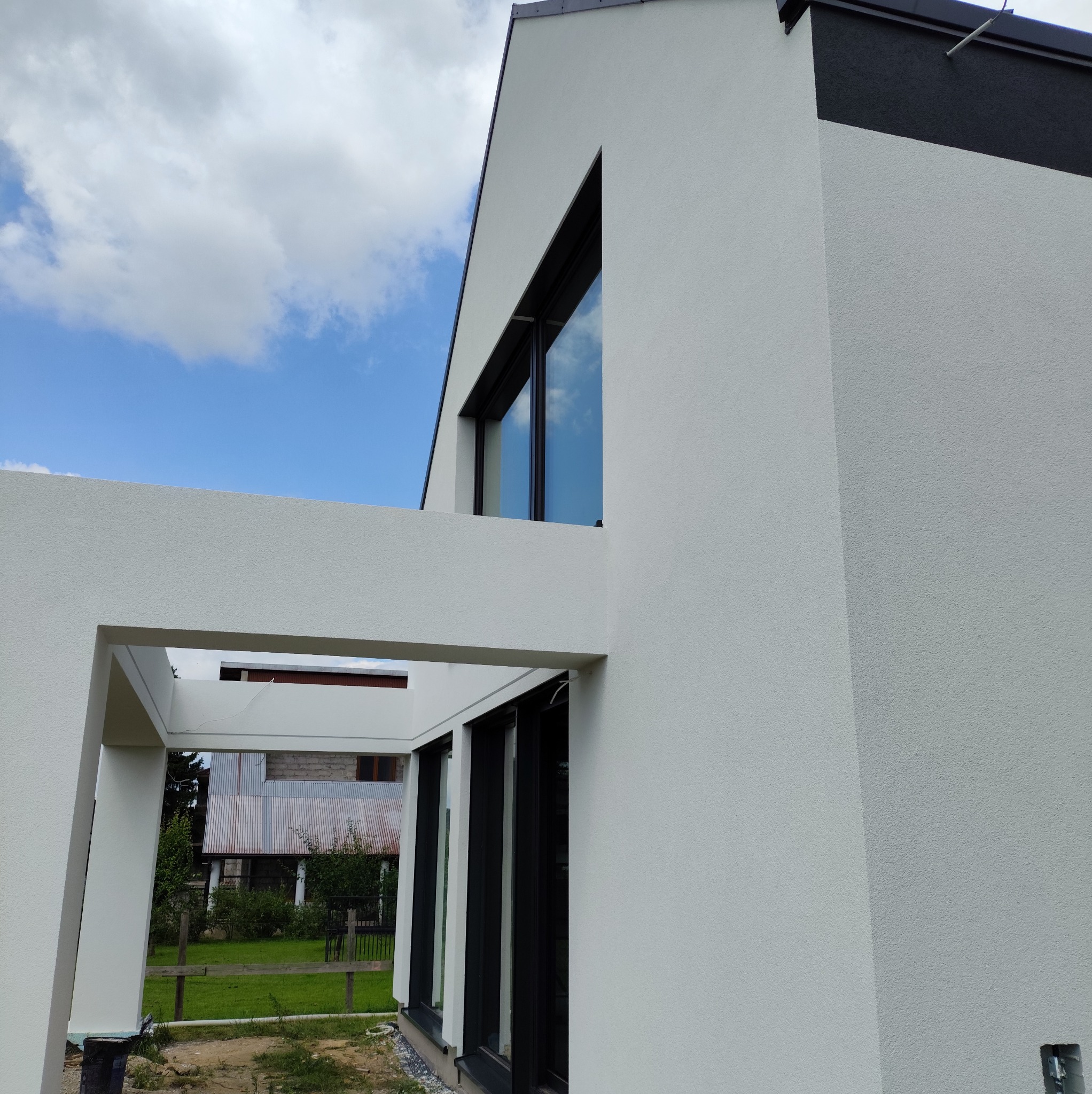 Nowoczesny dom z białą elewacją o fakturze baranka, czarne okna i fragment czarnego wykończenia dachu, w tle błękitne niebo z chmurami.