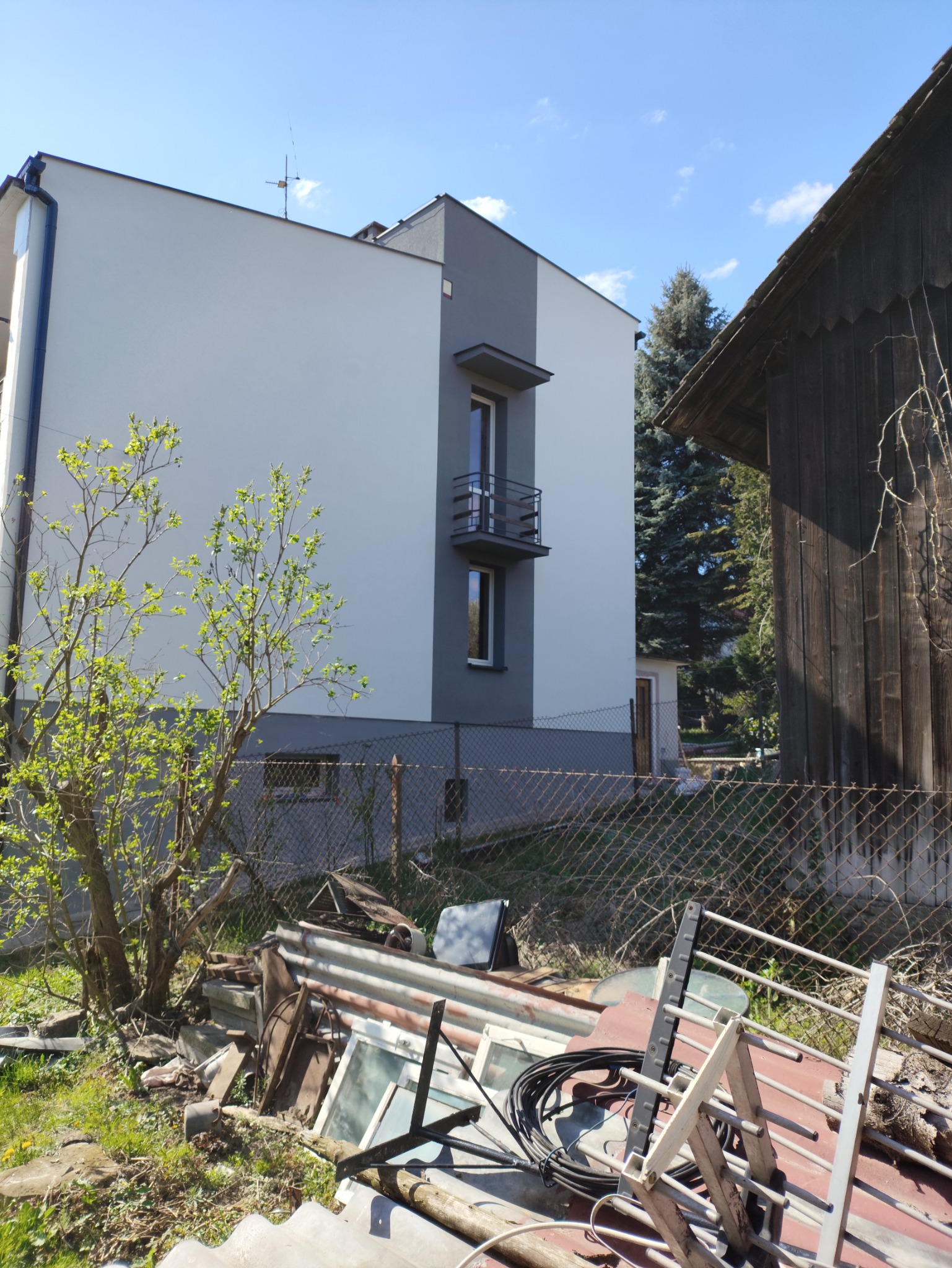 Dwukondygnacyjny budynek z nową, biało-szarą elewacją i balkonem, widziany zza ogrodzenia i sterty gruzu. W tle drewniany budynek i drzewa.