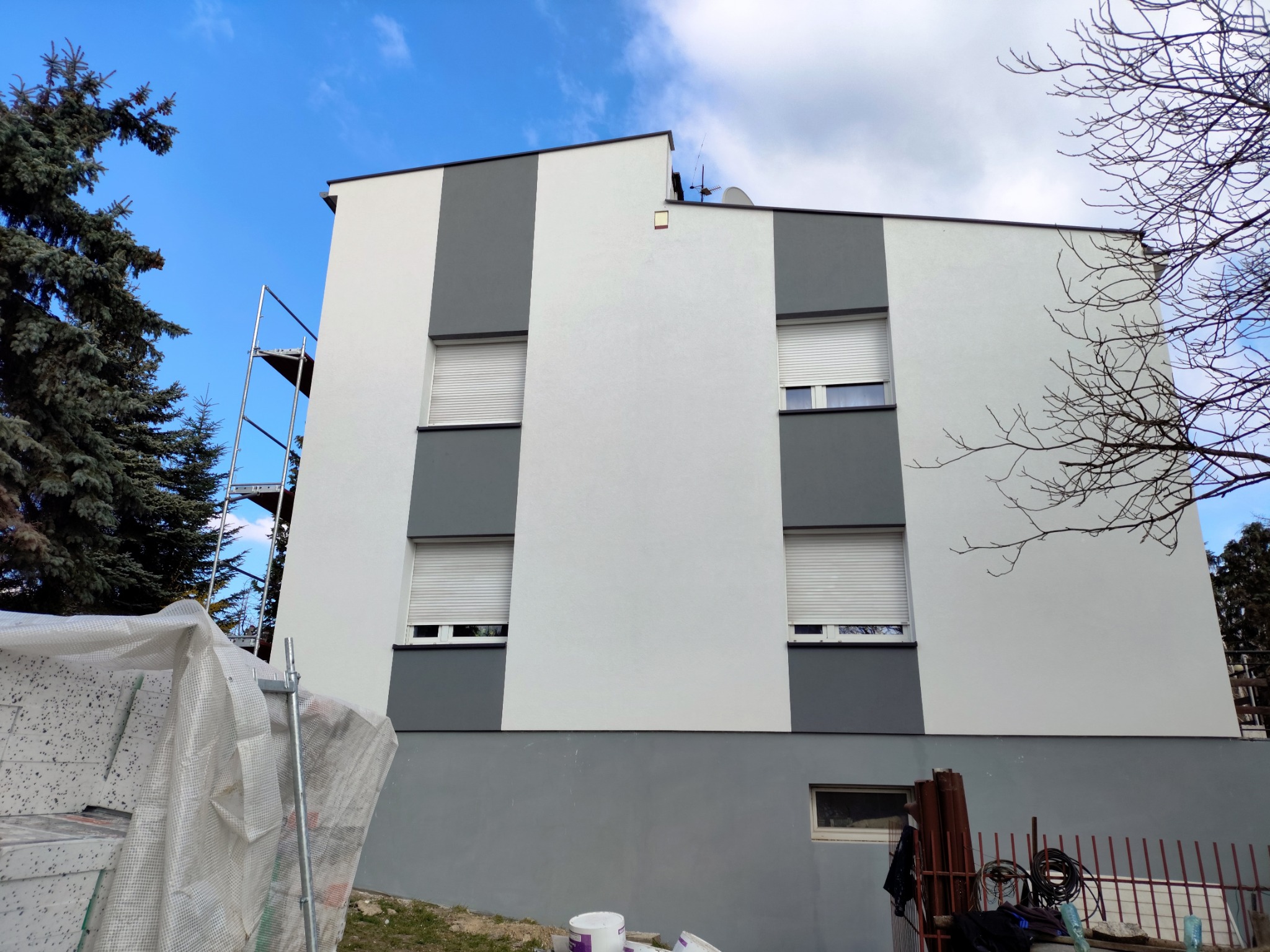 Dwukolorowa elewacja budynku z oknami i roletami, widziana z dołu. Na pierwszym planie rusztowanie i materiały budowlane. Błękitne niebo w tle.