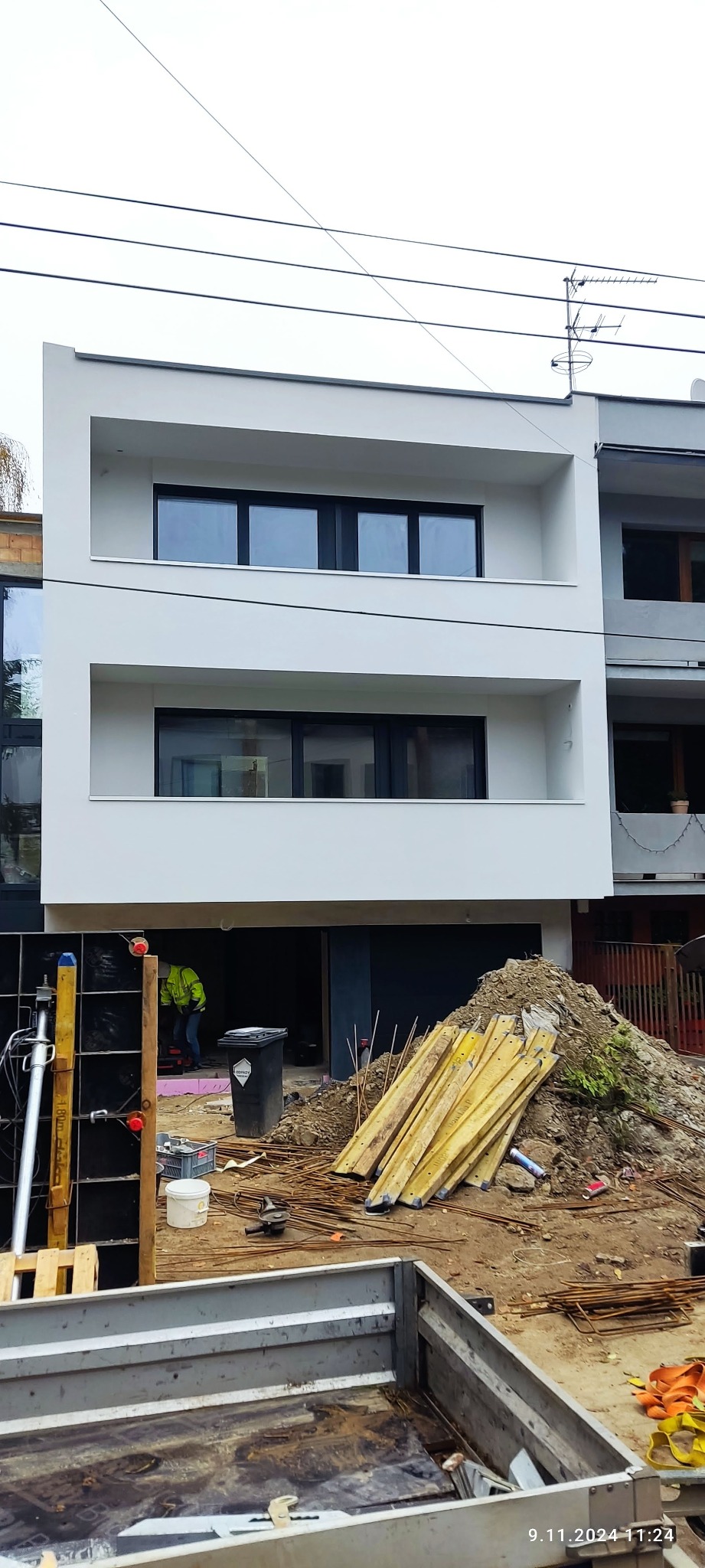 Nowoczesny, dwupiętrowy budynek z białą elewacją i czarnymi oknami, w trakcie prac wykończeniowych. Widoczne materiały budowlane i pracownik w odblaskowej kamizelce.