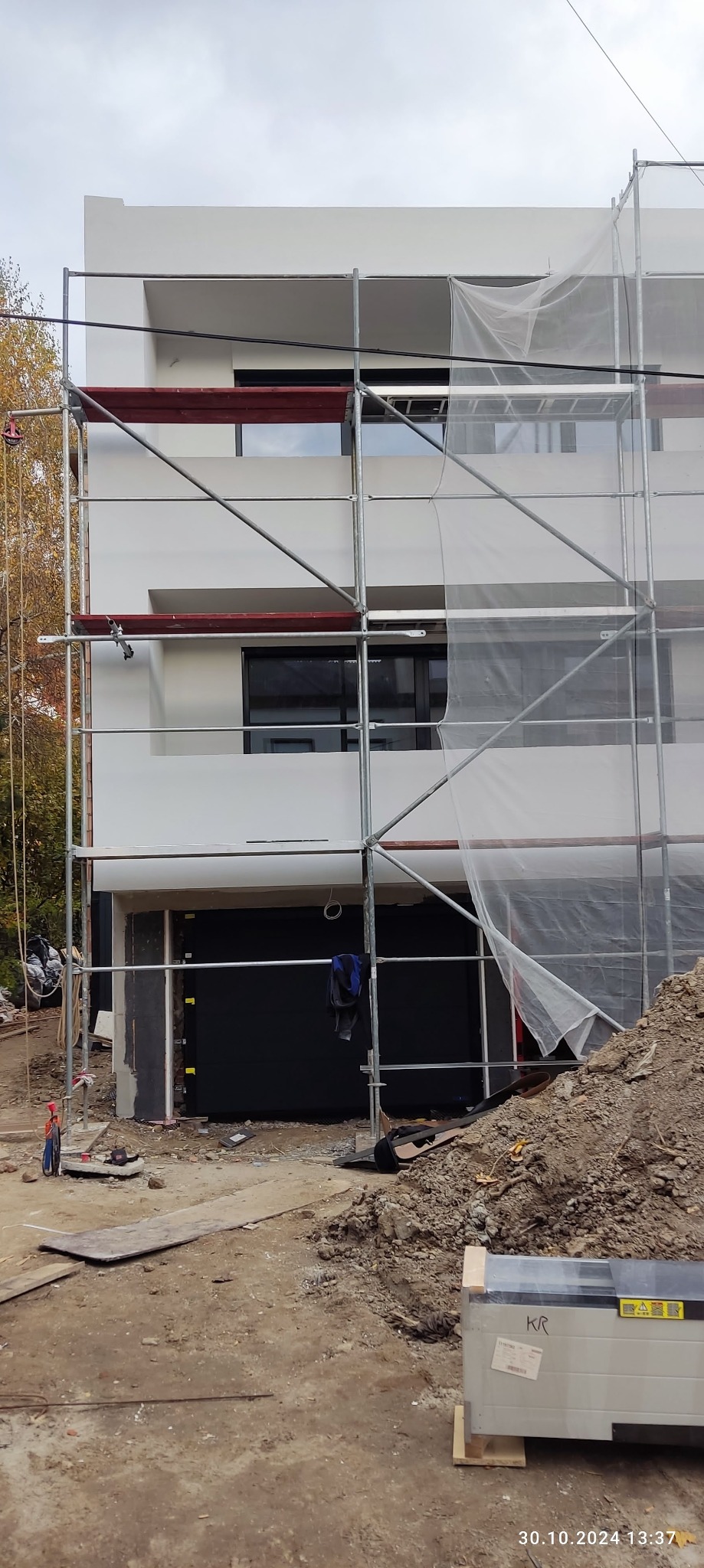 Dwupiętrowy budynek w trakcie prac elewacyjnych, z metalowymi rusztowaniami i częściowo osłonięty siatką. Widoczne narzędzia i materiały budowlane na ziemi.