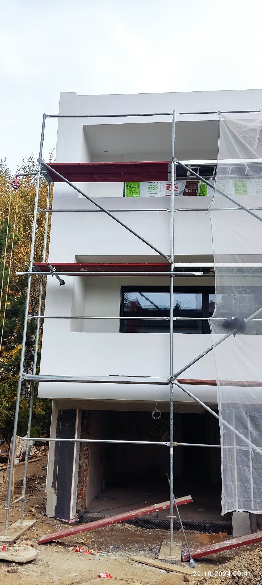 Dwupiętrowy budynek w trakcie prac elewacyjnych, z widocznymi rusztowaniami, częściowo zakryty folią ochronną, okna z czarną ramą i widoczne materiały izolacyjne.