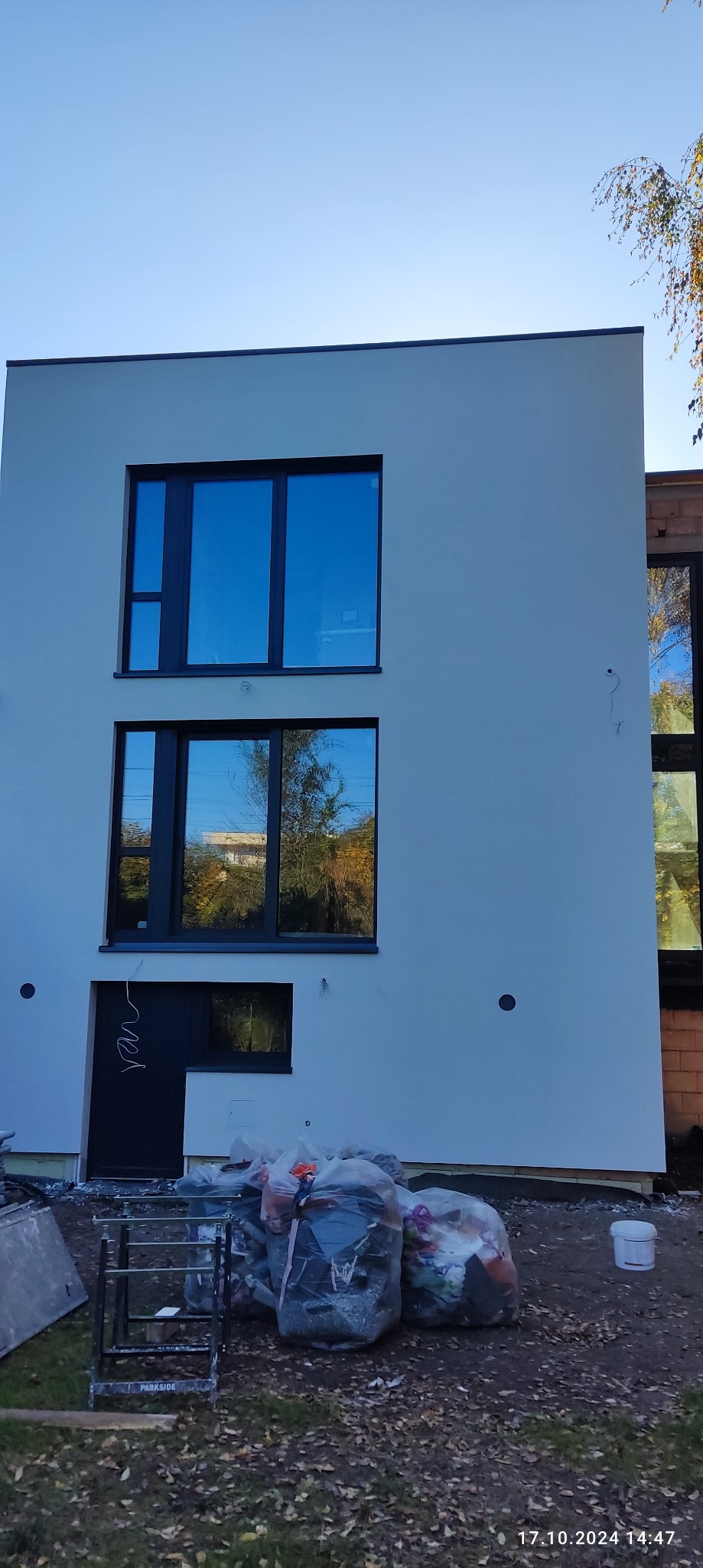 Biała elewacja dwukondygnacyjnego domu z czarnymi oknami, w których odbija się jesienne otoczenie. Na dole widoczne worki z odpadami budowlanymi i rusztowanie.