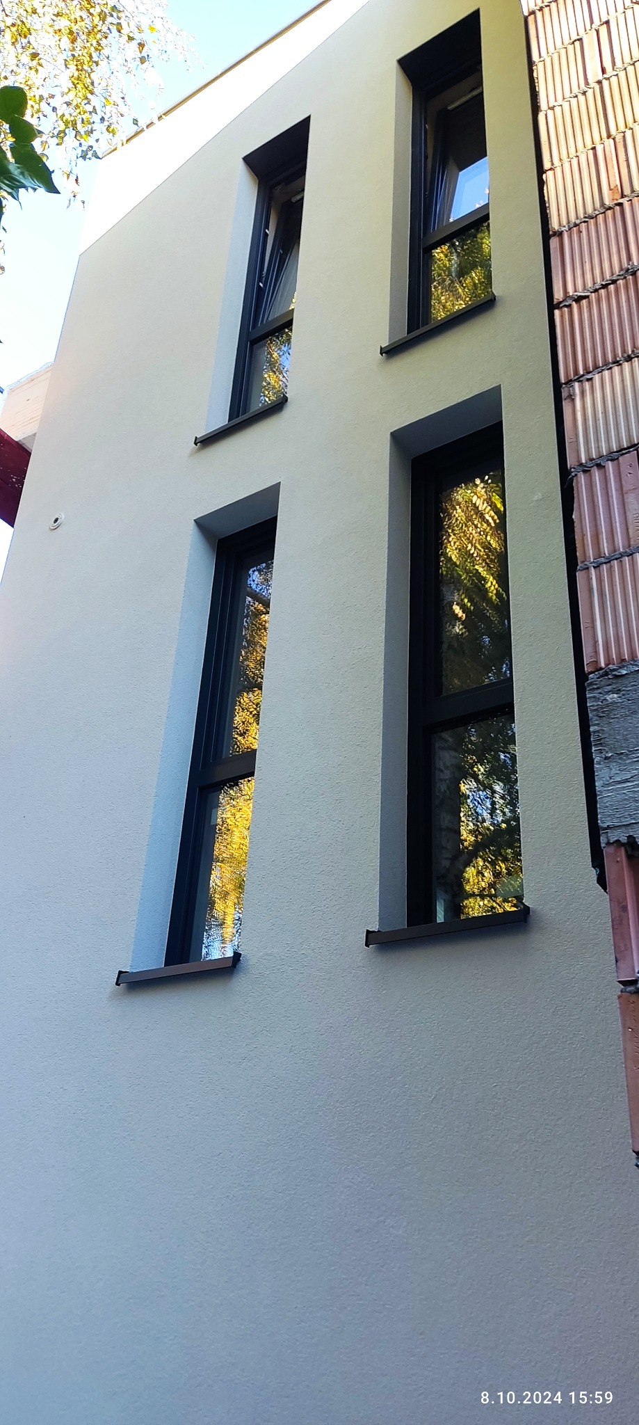 Jasna elewacja budynku z czterema oknami o czarnych ramach. Po prawej stronie widoczny fragment ściany z cegły. W oknach odbijają się drzewa.