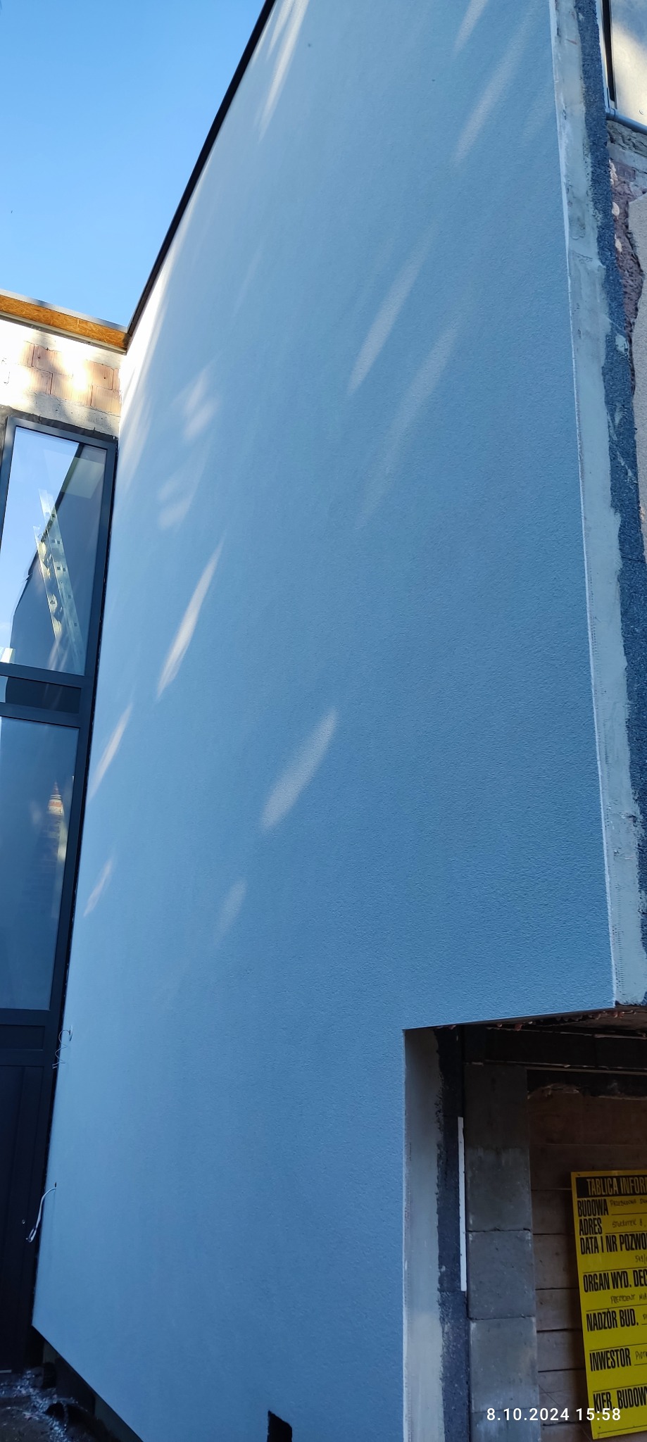 Ściana budynku po tynkowaniu, pomalowana na niebiesko, z widocznymi refleksami światła. W tle okno i fragment niezakończonej elewacji z tablicą informacyjną budowy.