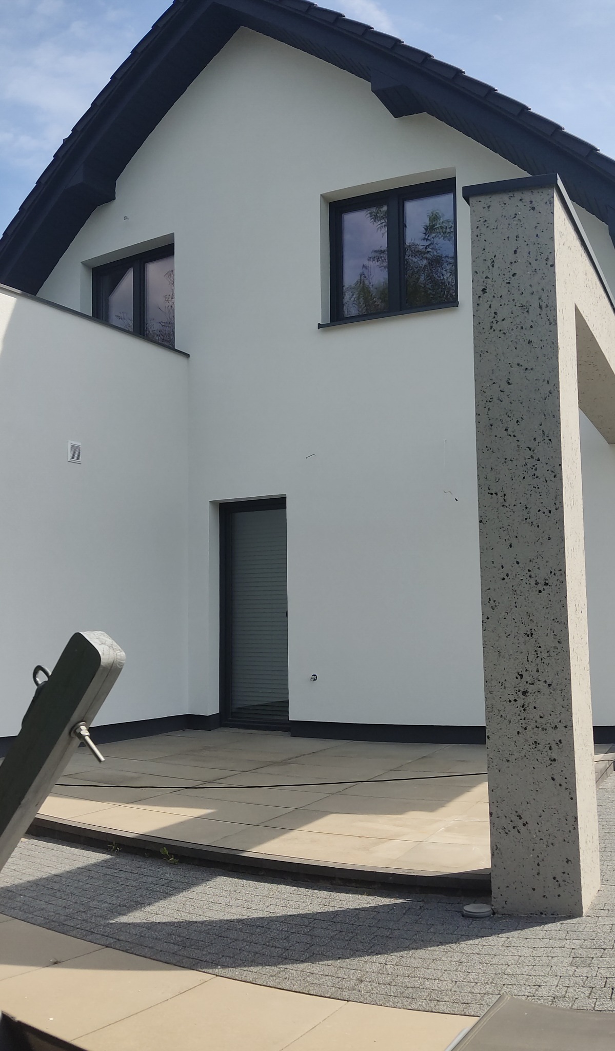 Biała elewacja domu z czarną stolarką okienną i dachem. Widoczny fragment tarasu wyłożonego jasnymi płytami i betonowa kolumna.